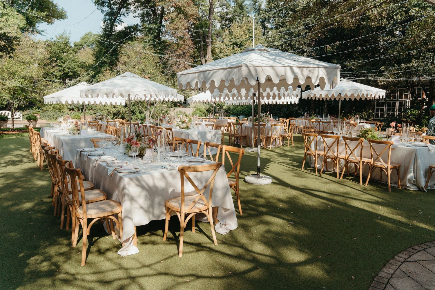 Elegant outdoor wedding reception with white striped umbrellas, wooden cross-back chairs, and long linen-draped tables on green lawn.