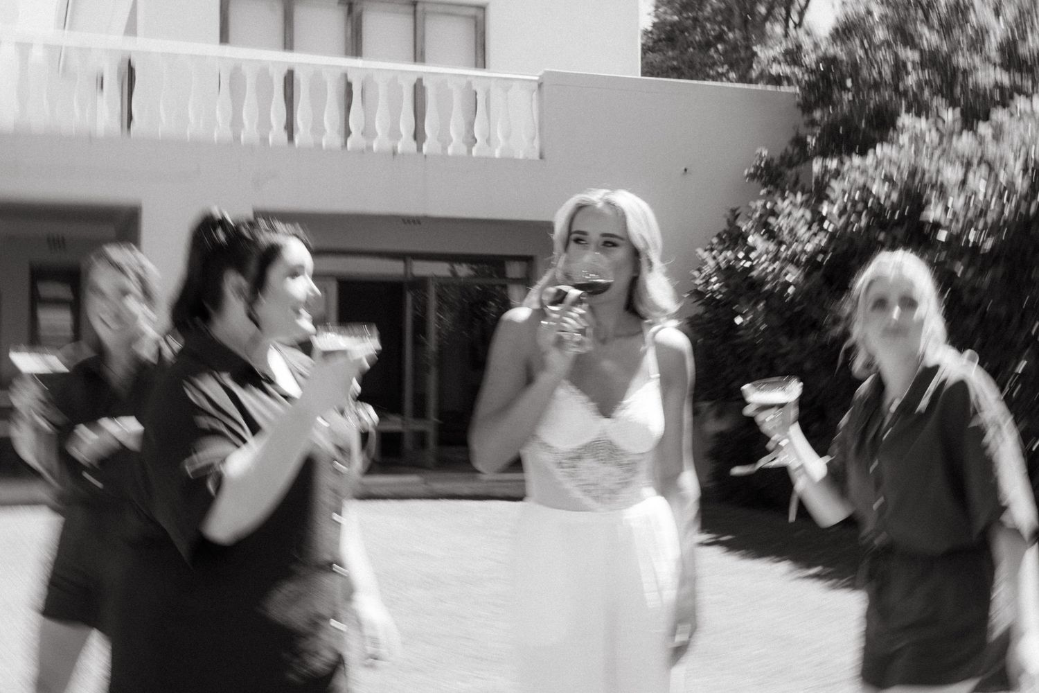 Black and white photo of a bride in white dress celebrating with friends outdoors near a building, all holding drinks.
