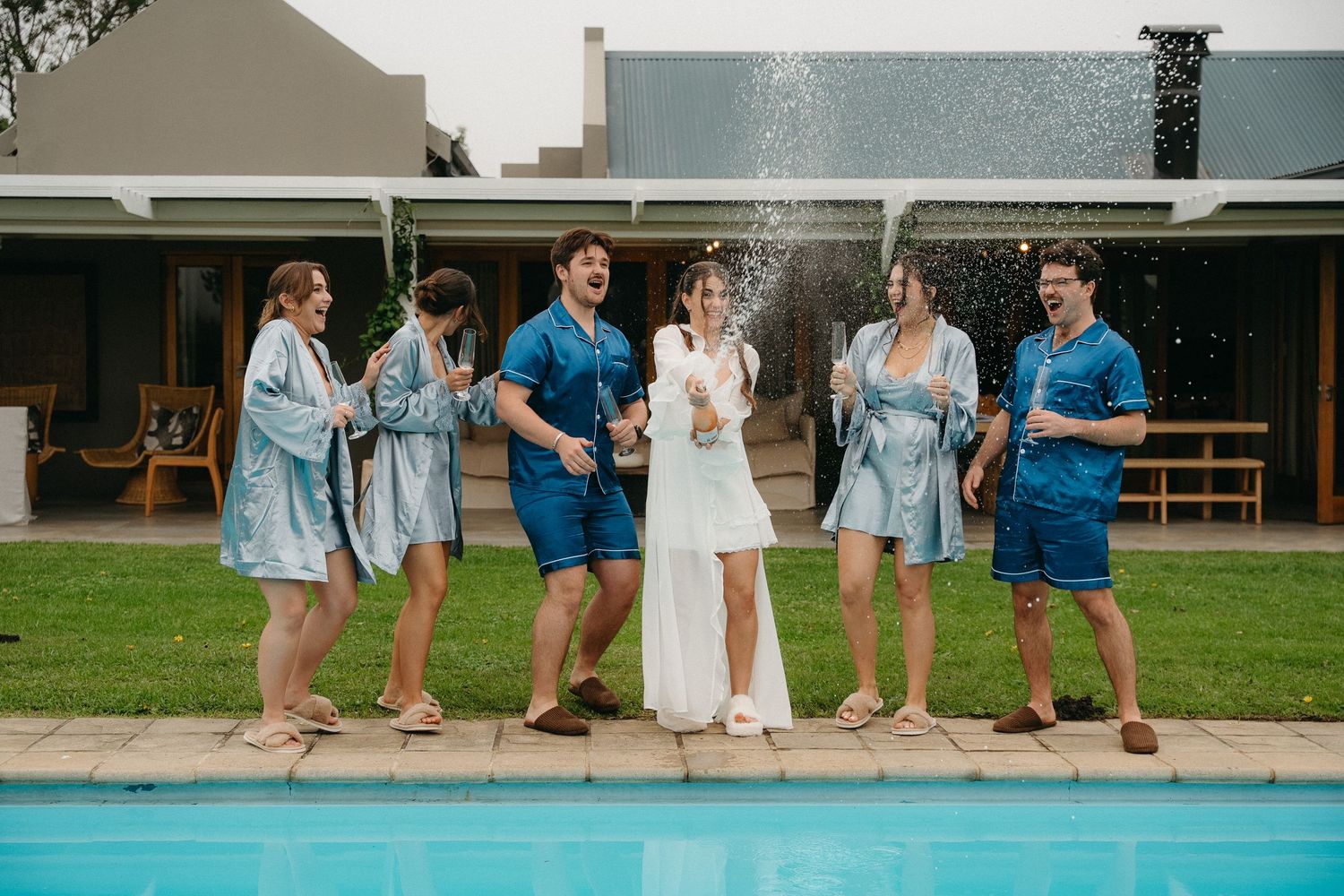 Bride and wedding party celebrating with champagne by a pool, wearing matching blue robes outdoors.