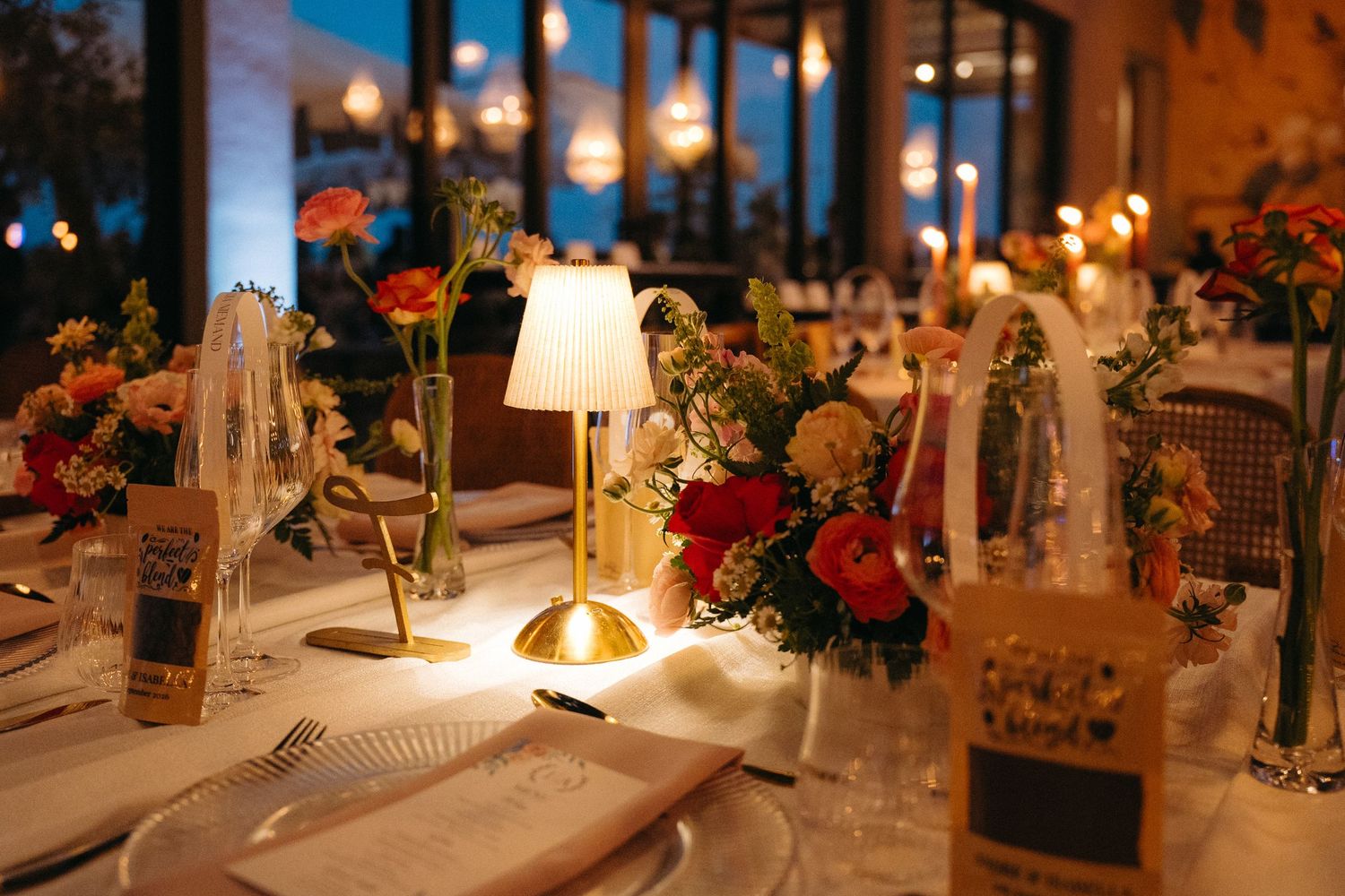 Elegant wedding reception table with gold lamp, colorful floral centerpiece, and warm string lights in background.
