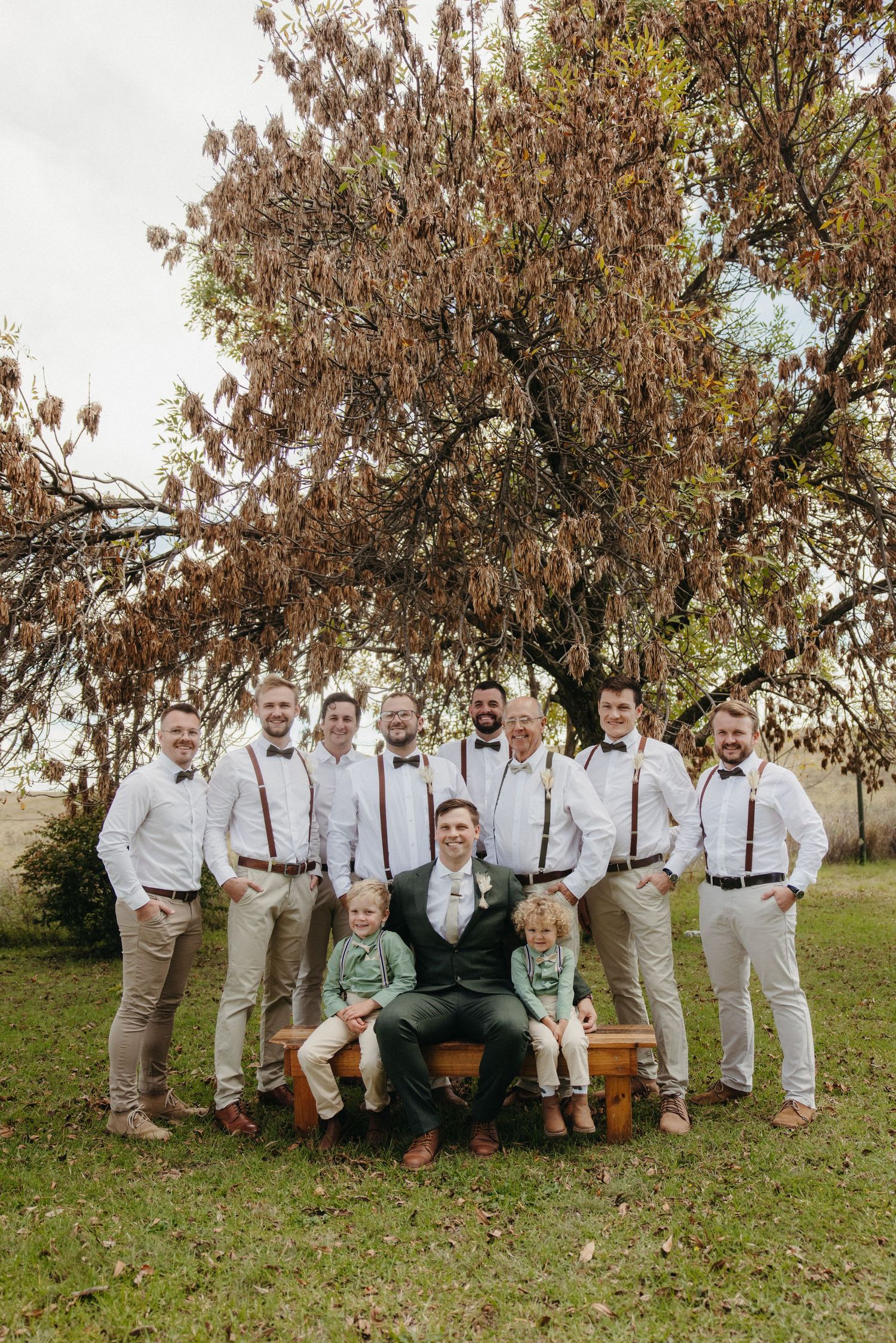 Groom and groomsmen in white shirts with suspenders pose outdoors near a tree, with two young children seated on a bench.