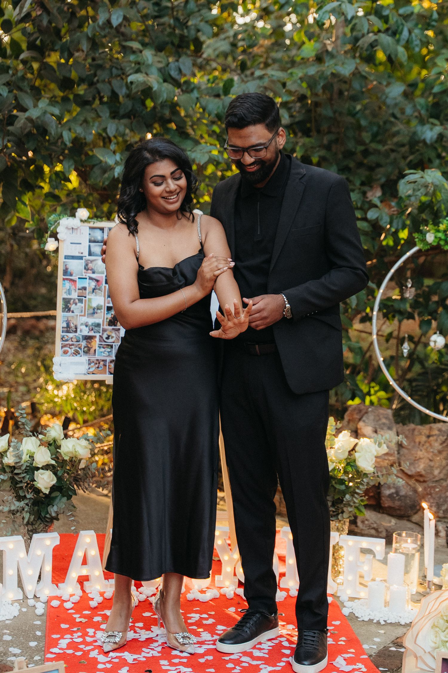 Couple in matching black outfits sharing a romantic ring exchange moment outdoors at an elegant proposal setup.