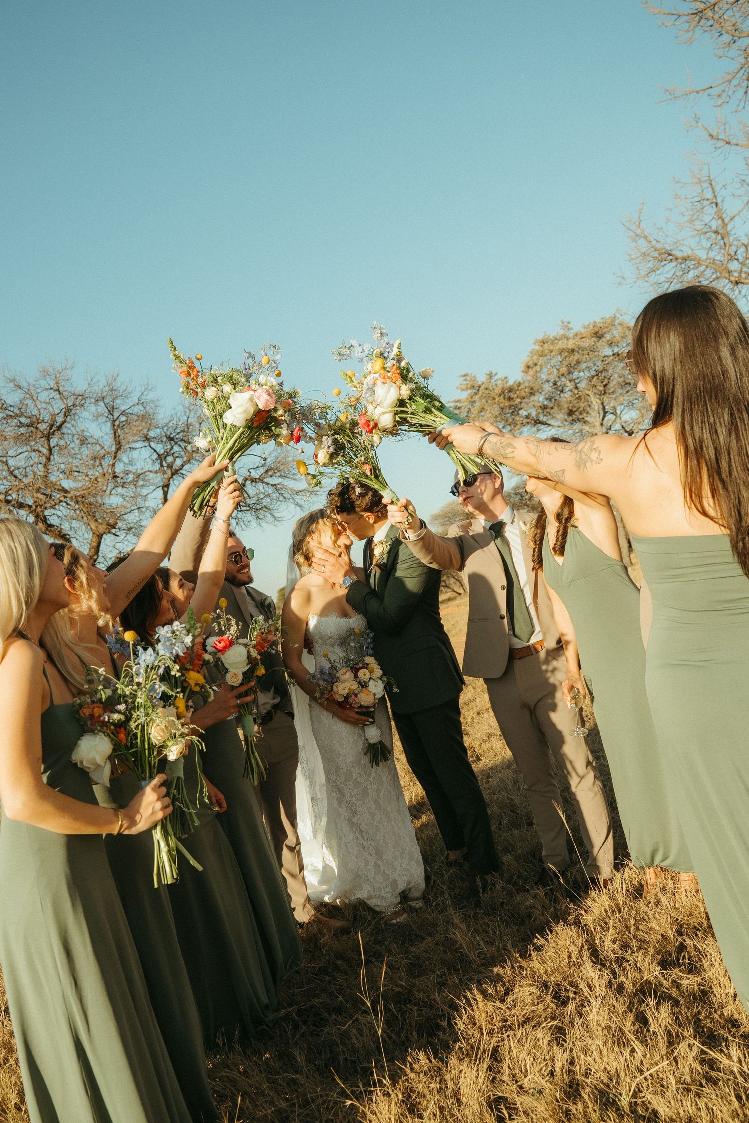 Bride and groom kiss under an arch of colorful bouquets held by bridesmaids in sage green dresses at Monate Game Lodge.
