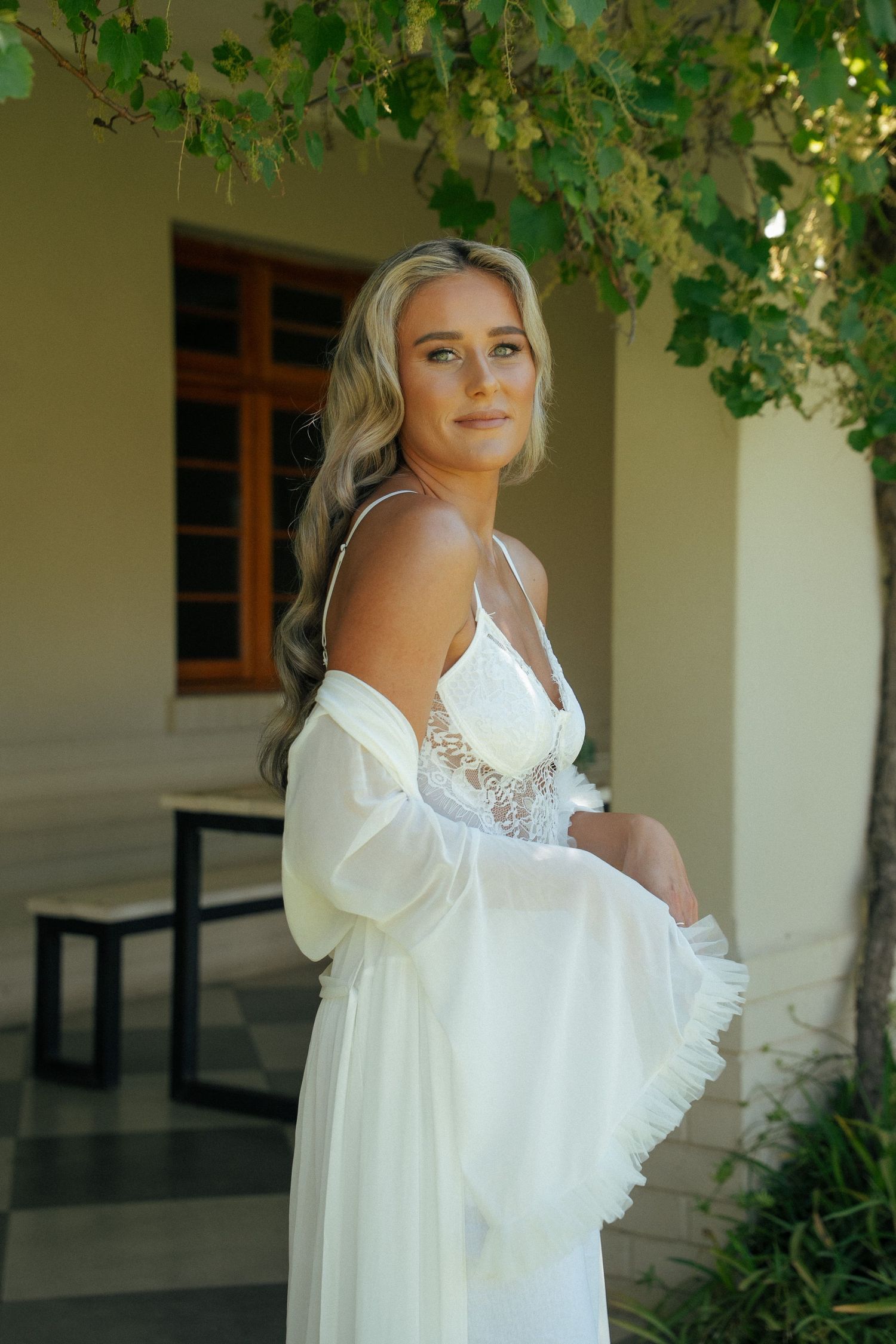 Bride with wavy blonde hair wears a white lace bridal slip dress and flowing chiffon robe, posing outdoors beneath leafy vines at Duvon.