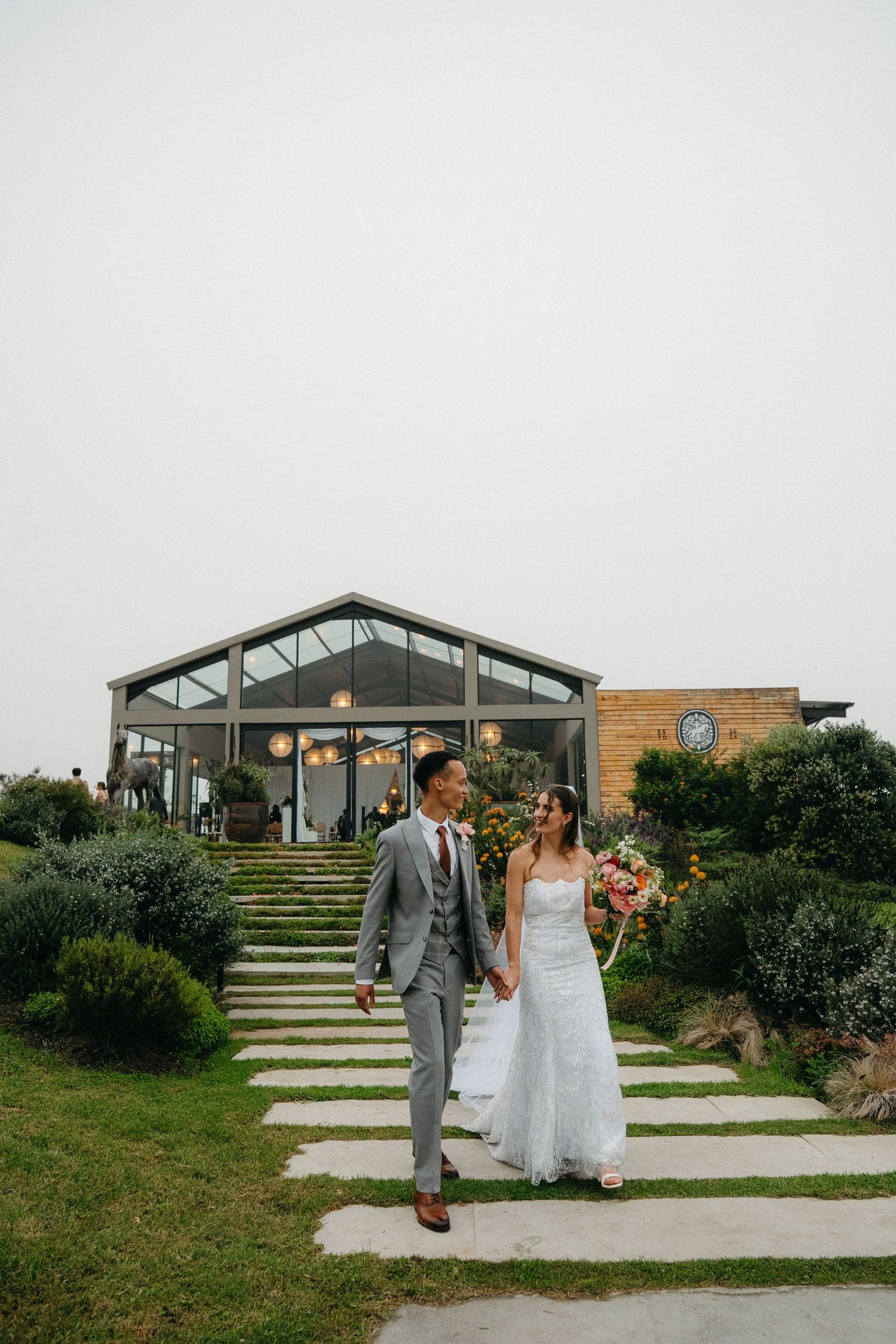 A bride and groom walk hand-in-hand along a stone path outside a glass wedding venue surrounded by lush gardens.