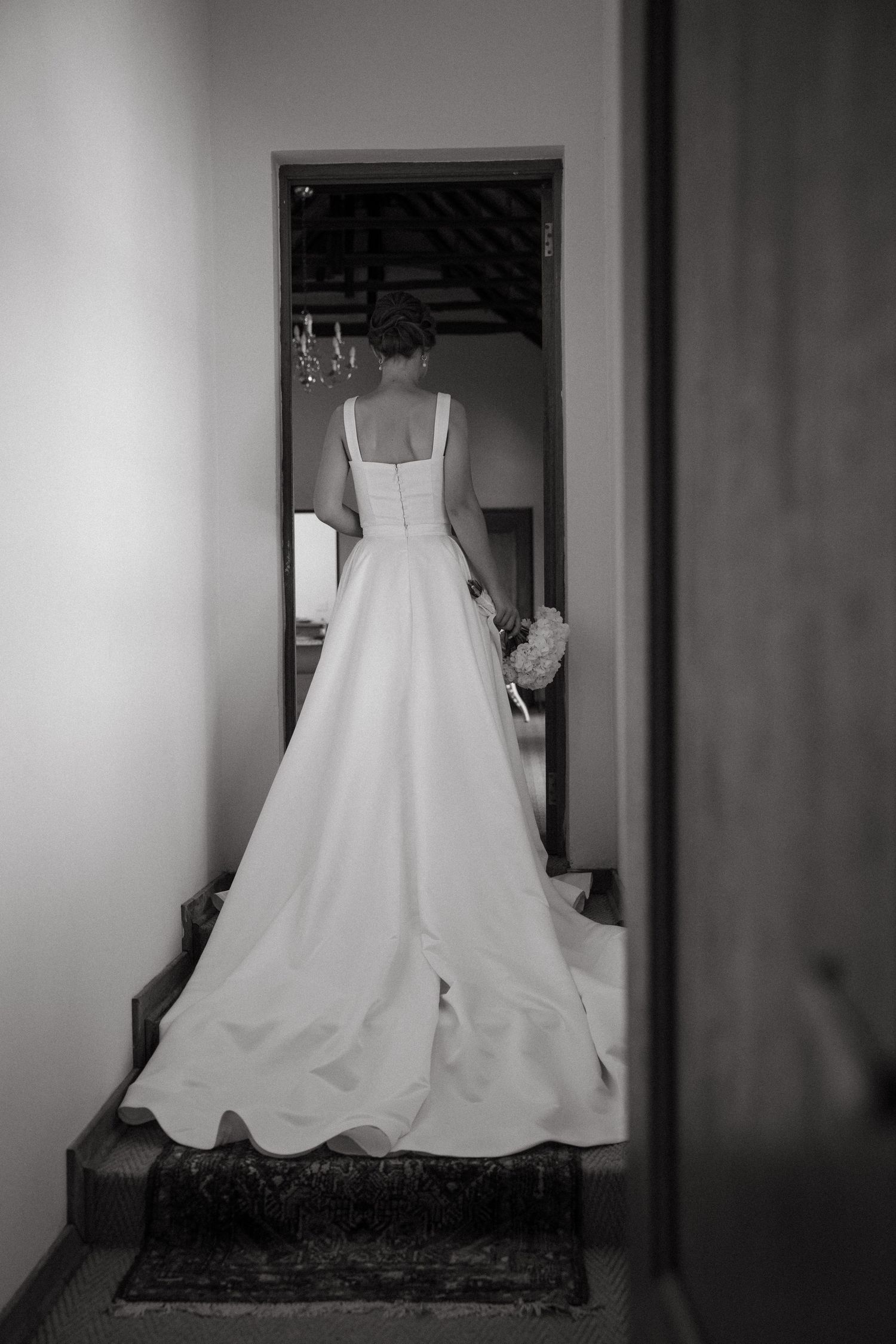 Black and white photo of a bride in an elegant white wedding gown standing in a doorway, viewed from behind.