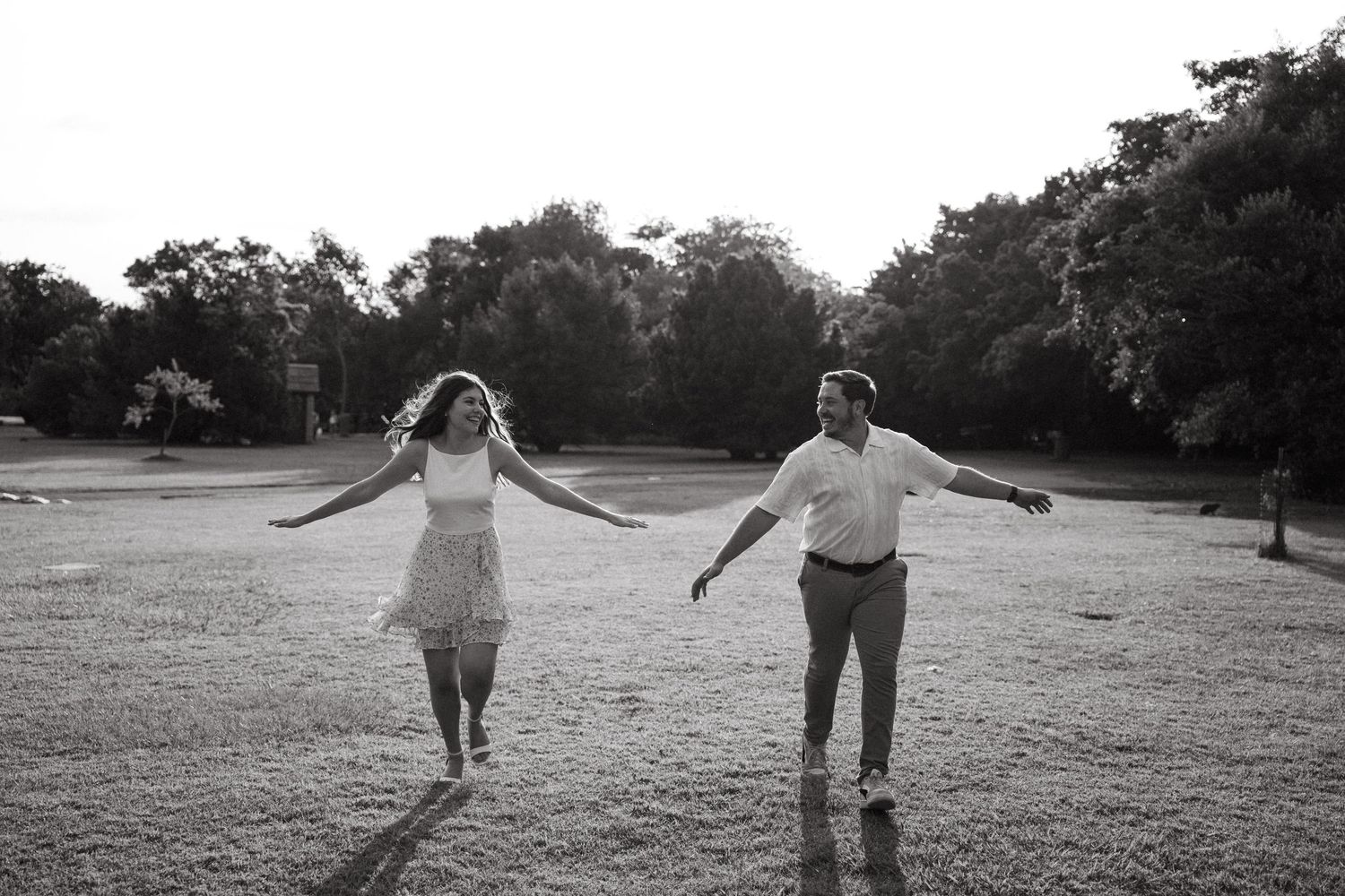 Black and white photo of a joyful couple holding hands and spinning in an open park surrounded by trees.