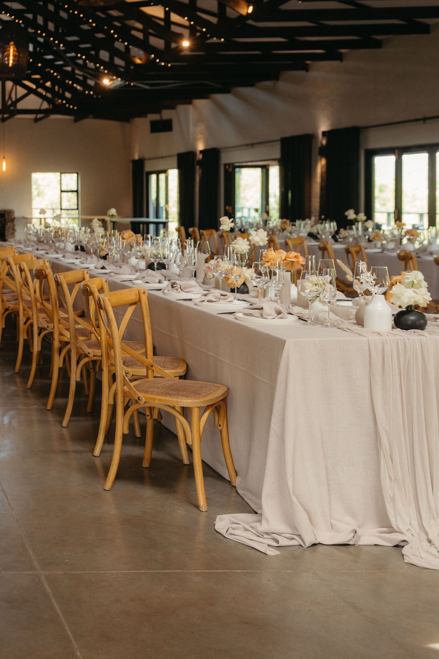 Long banquet tables with white linens and wooden cross-back chairs set up in an elegant reception venue with exposed beams.