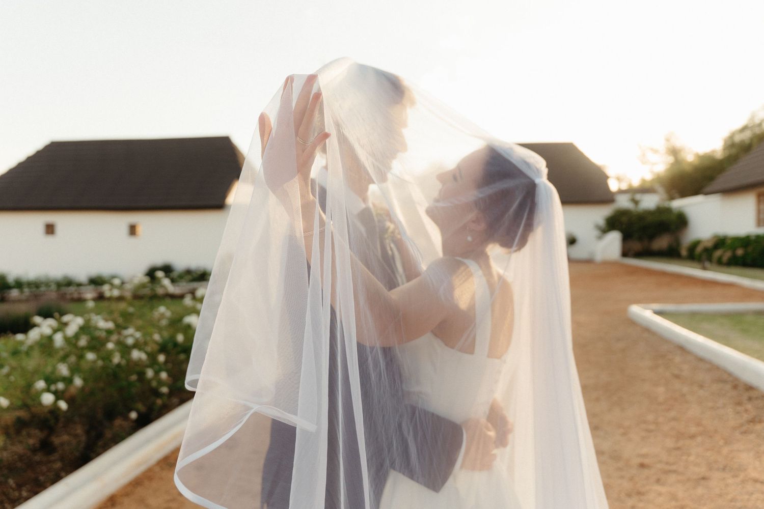 A couple in wedding attire embraces during sunset at a beachside venue.