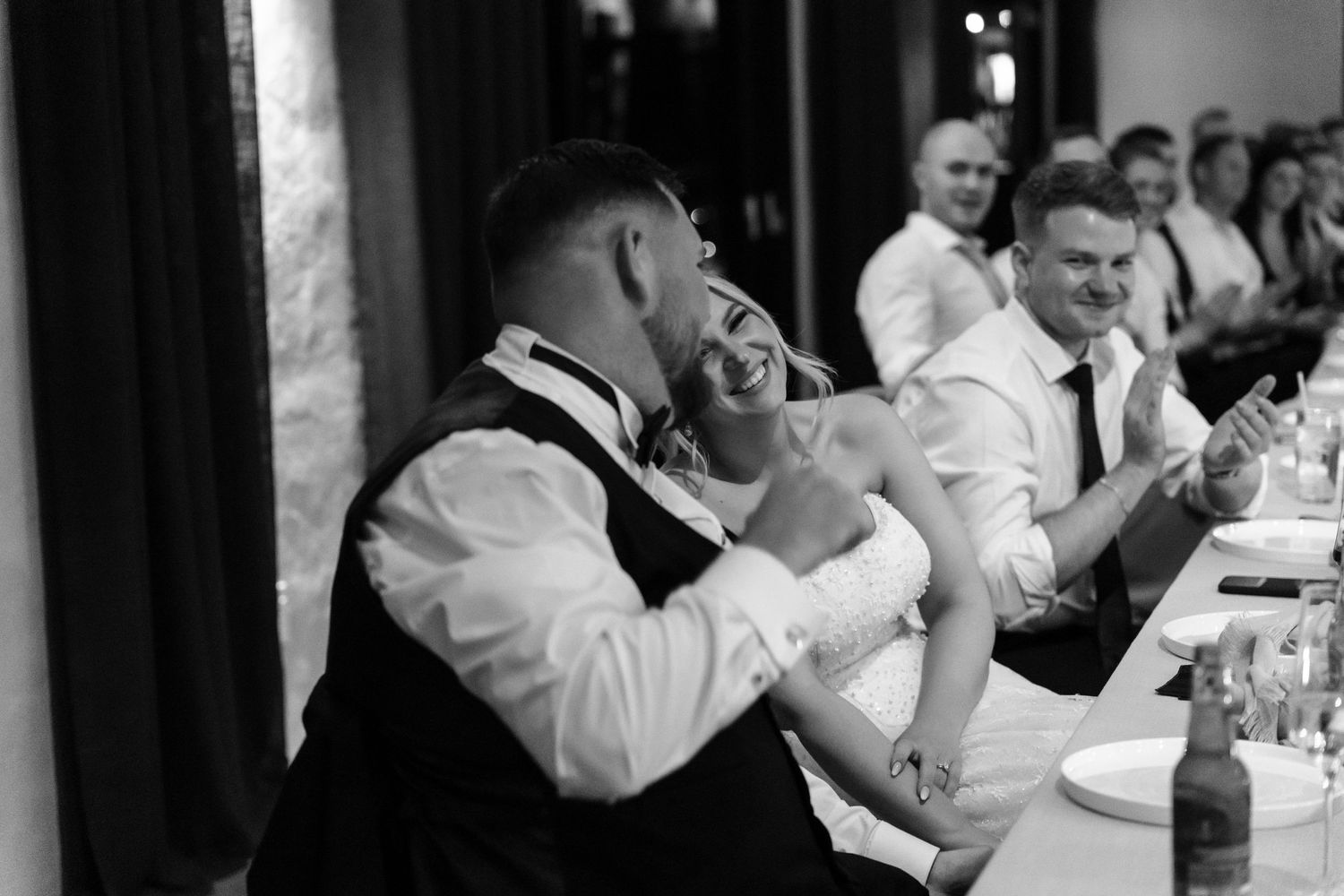 Wedding guests laugh and smile while seated at round tables during an elegant reception in black and white.