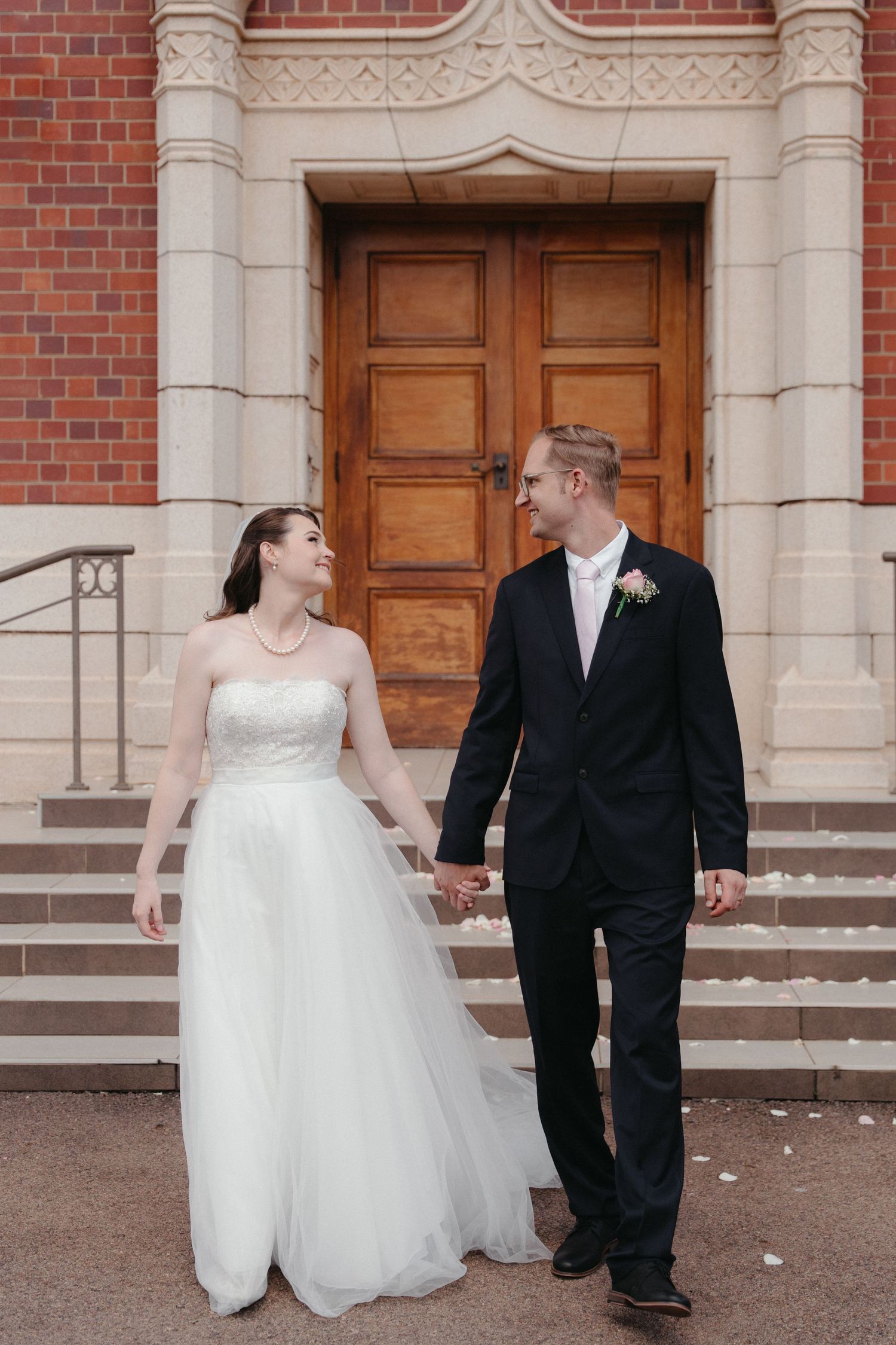 A couple in wedding attire holds hands while walking up brick and stone steps outside a historic building.