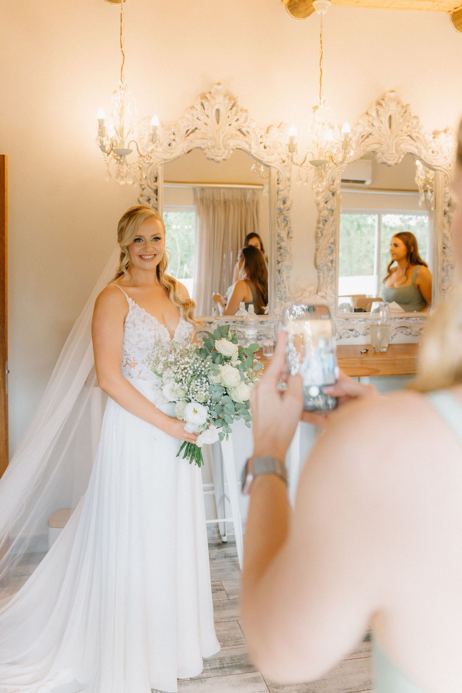 Bride in white gown getting ready in ornate bridal suite with elegant chandeliers and mirrors.