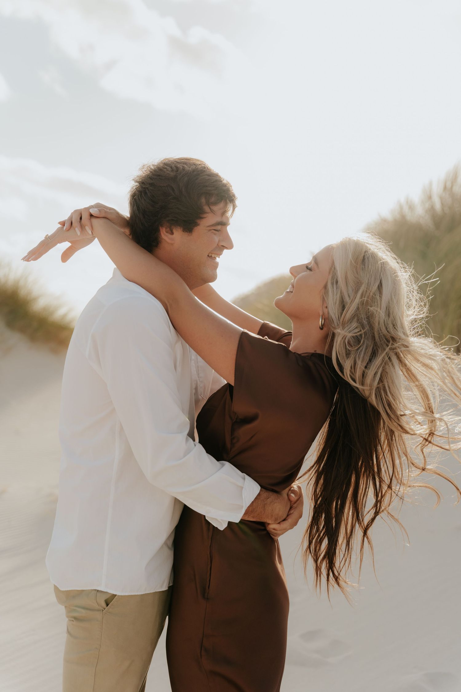 A joyful couple embraces on a sandy beach, laughing together, woman in brown dress with flowing hair.