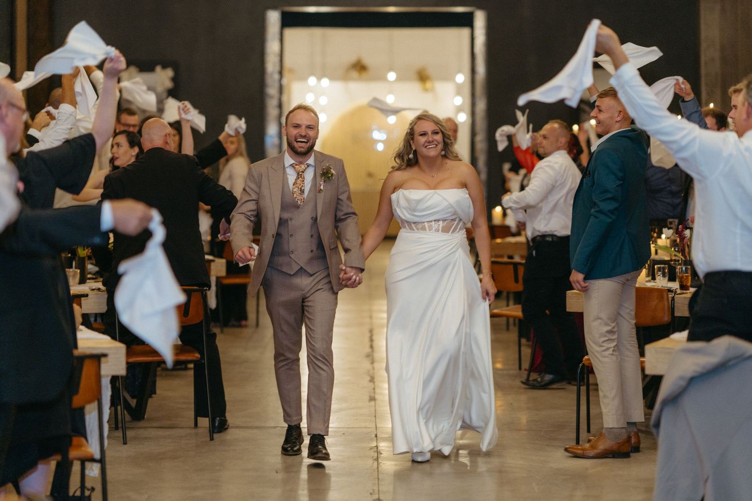 Wedding celebration scene with guests waving white napkins as couple walks through reception venue.