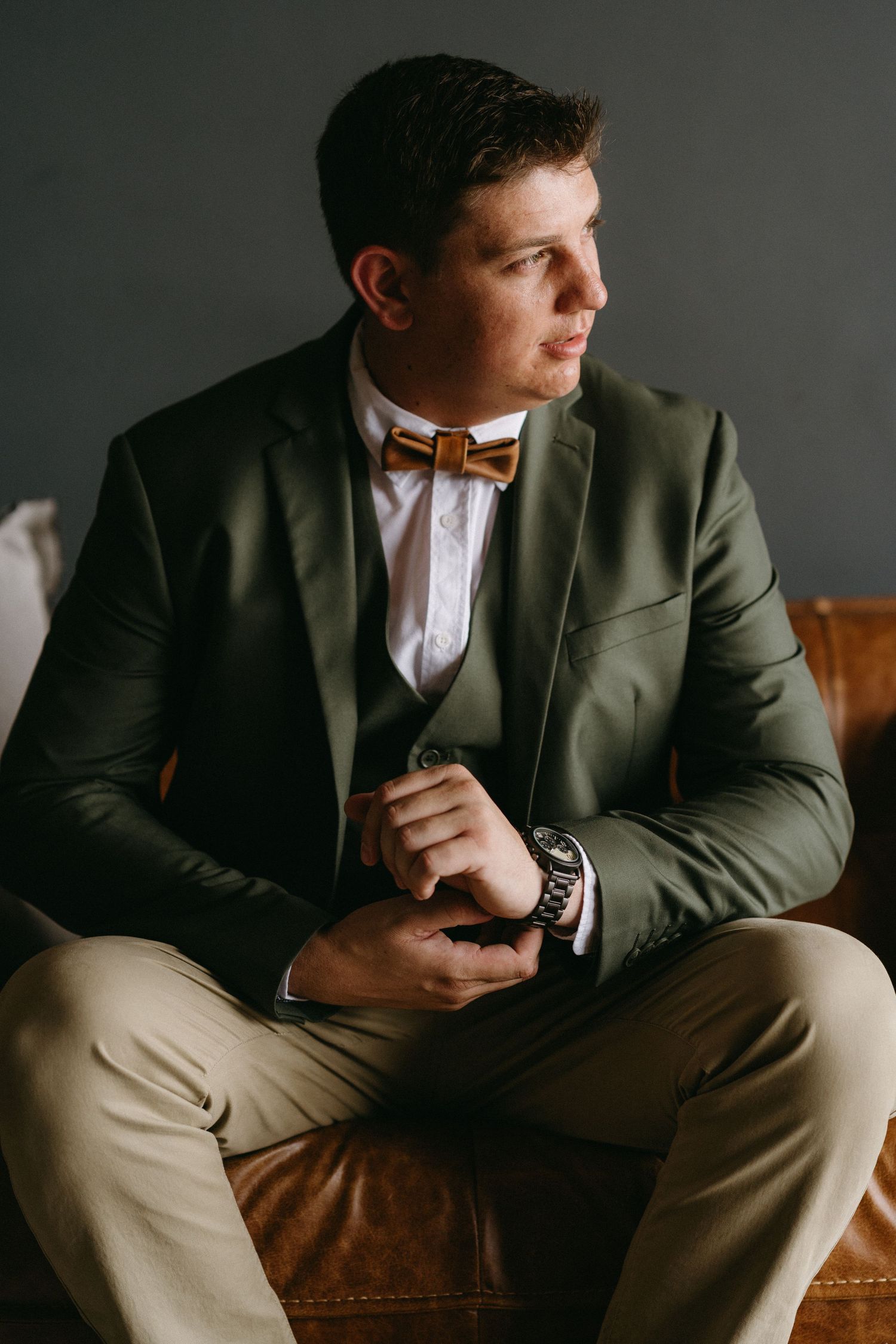 Stylish green blazer and khaki pants outfit paired with brown bow tie and wristwatch on leather sofa.
