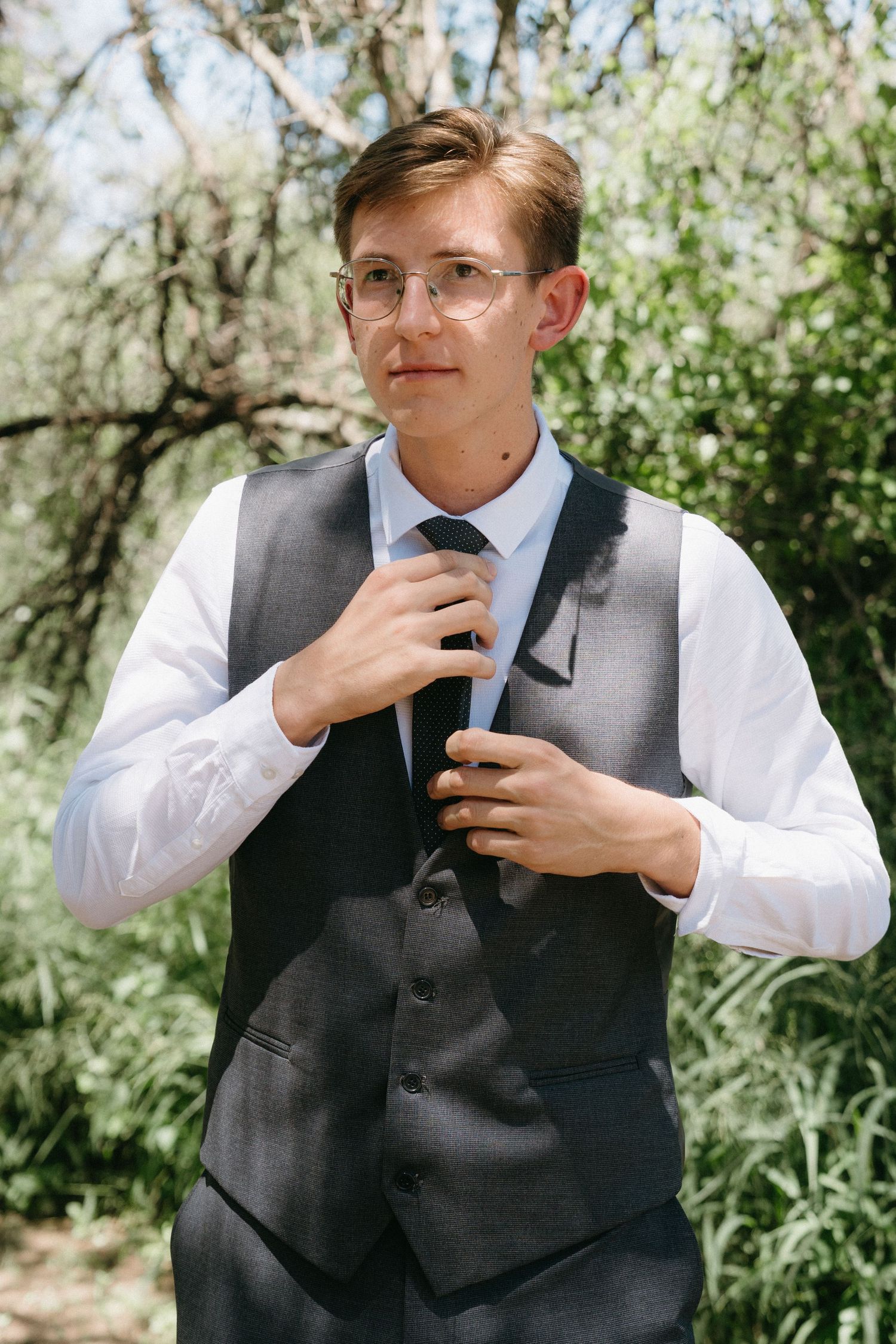 A young man in formal attire adjusts his tie while standing in front of blooming spring foliage.
