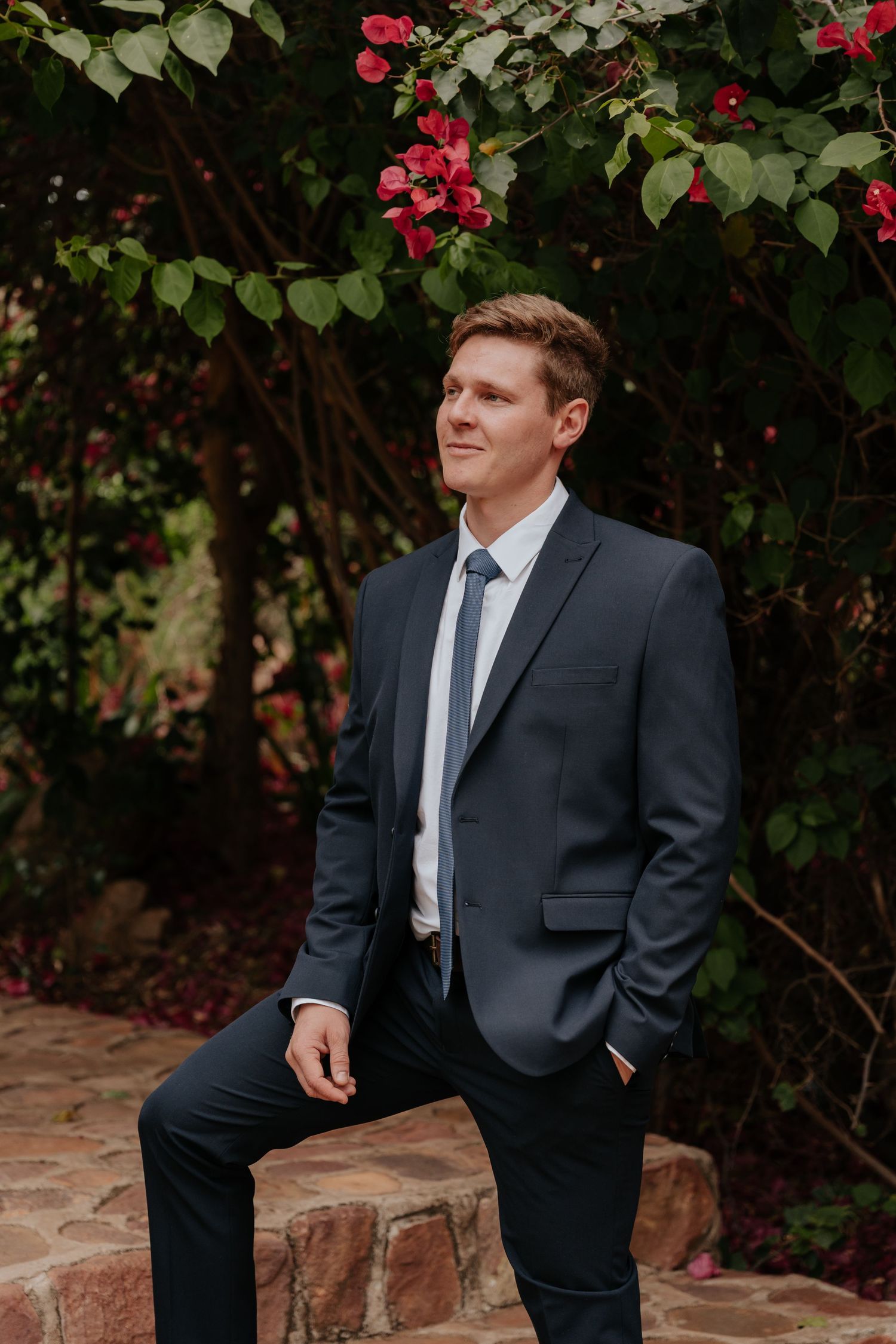 Man in dark suit sitting on brick ledge with red flowering bushes in background.