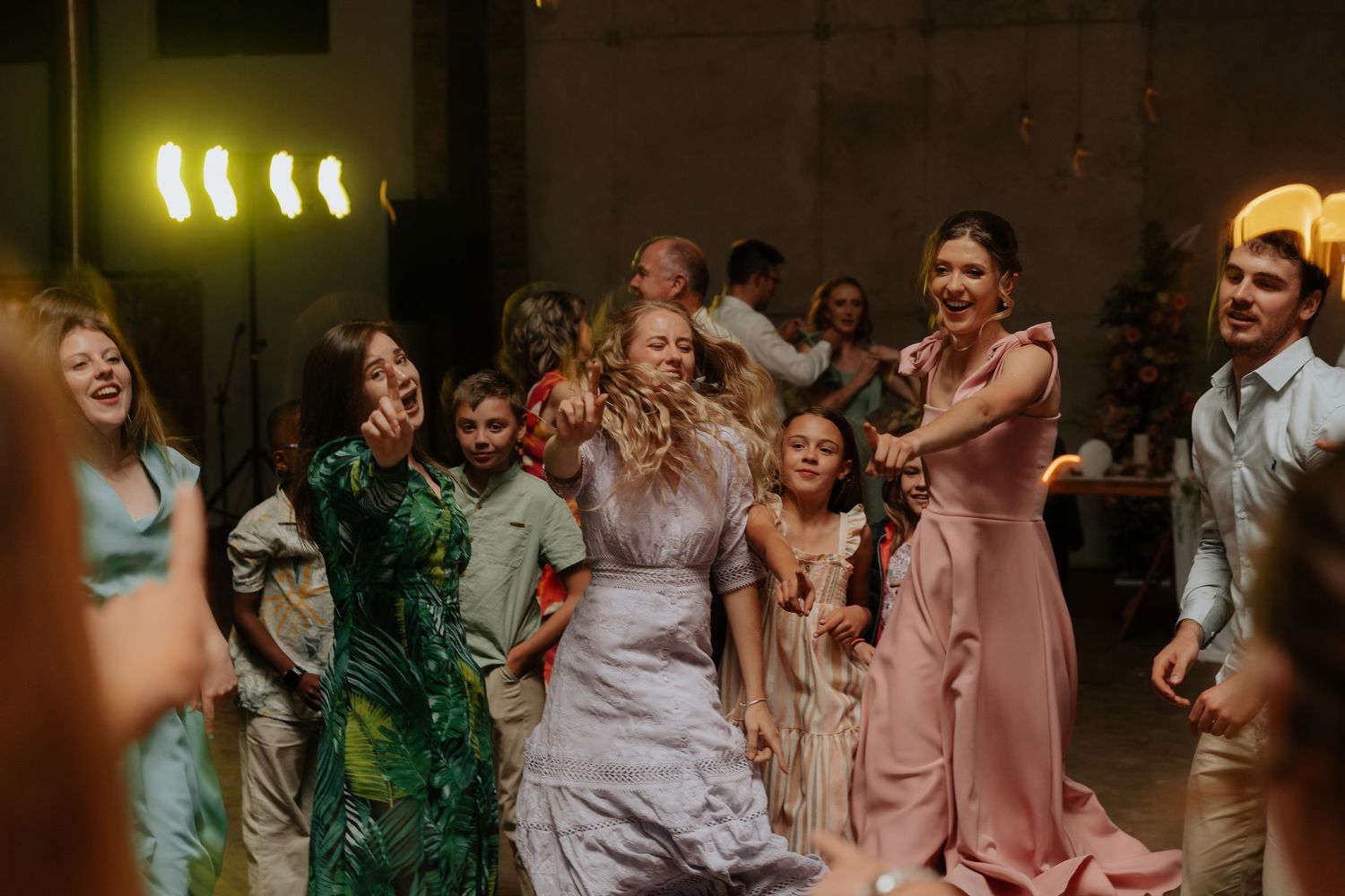 Wedding party members dancing in pink and white attire under warm lighting at an indoor reception venue.