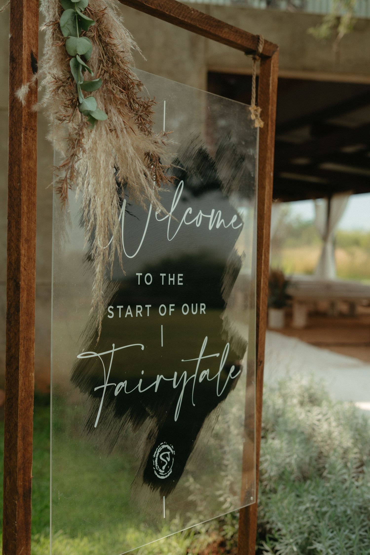 Rustic welcome sign with 'Welcome to the Start of our Fairytale' written in elegant script on glass with dried pampas grass decor.