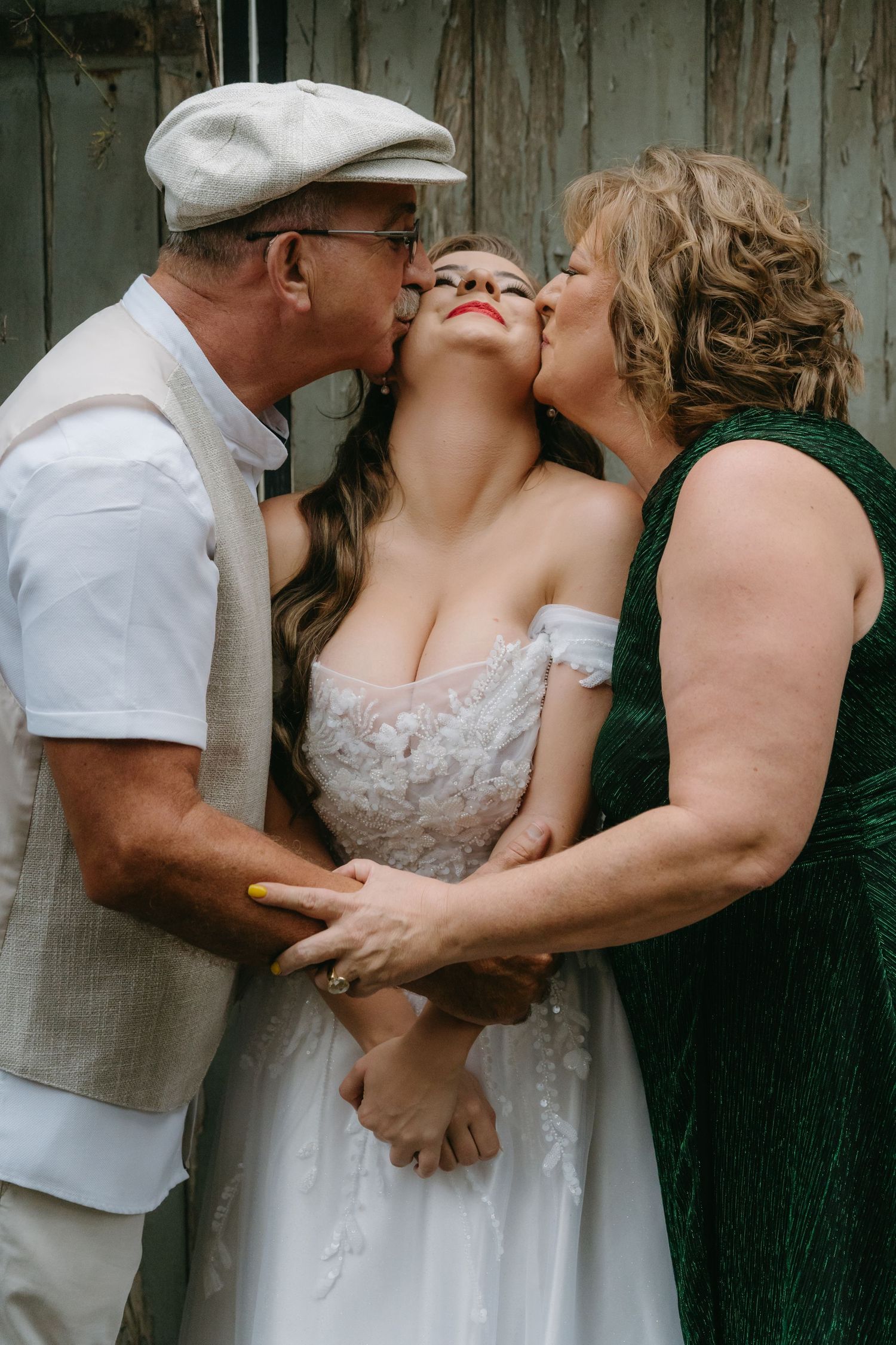 Parents giving loving kiss to bride in white lace dress against weathered wood backdrop.