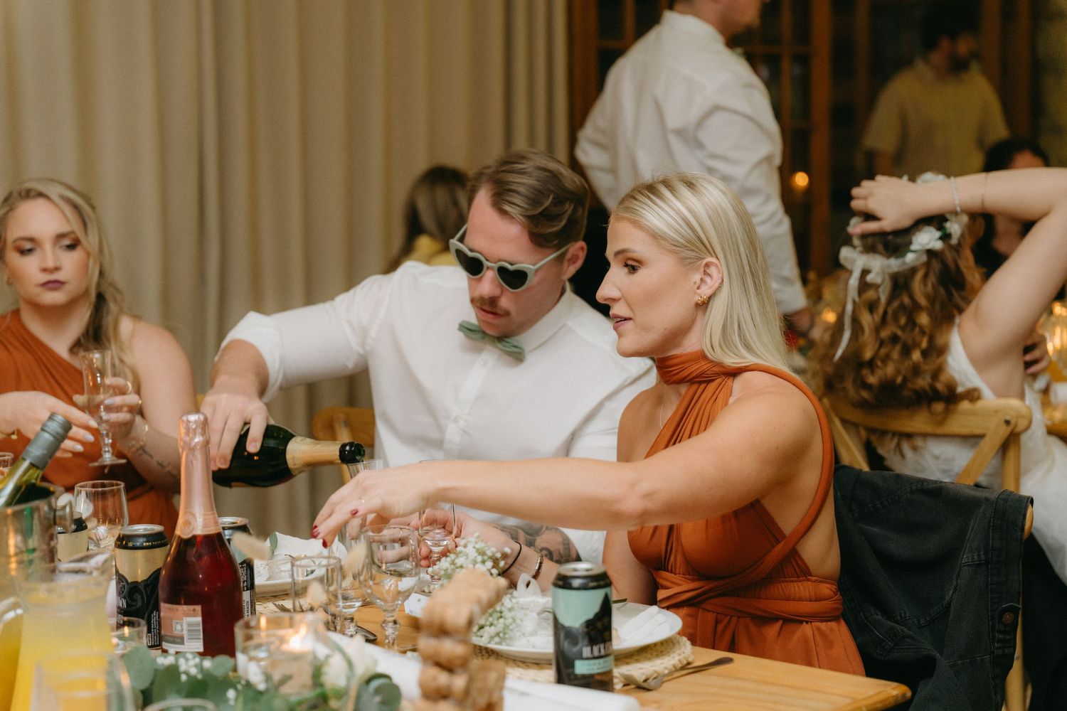Wedding reception guests enjoying dinner and conversation at elegantly decorated tables with warm lighting.