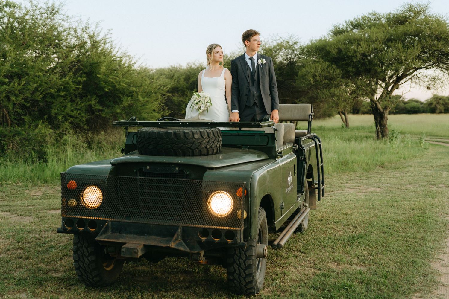 A couple on their wedding day stands in a classic dark green Land Rover with its headlights on in a rustic outdoor setting.