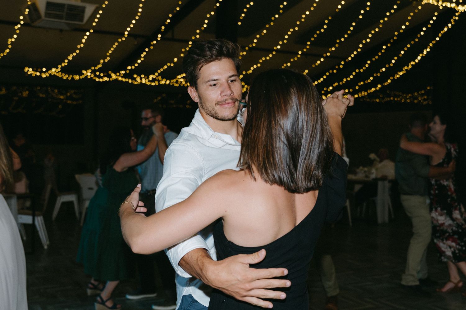 A couple slow dances together under glowing string lights at an evening reception.
