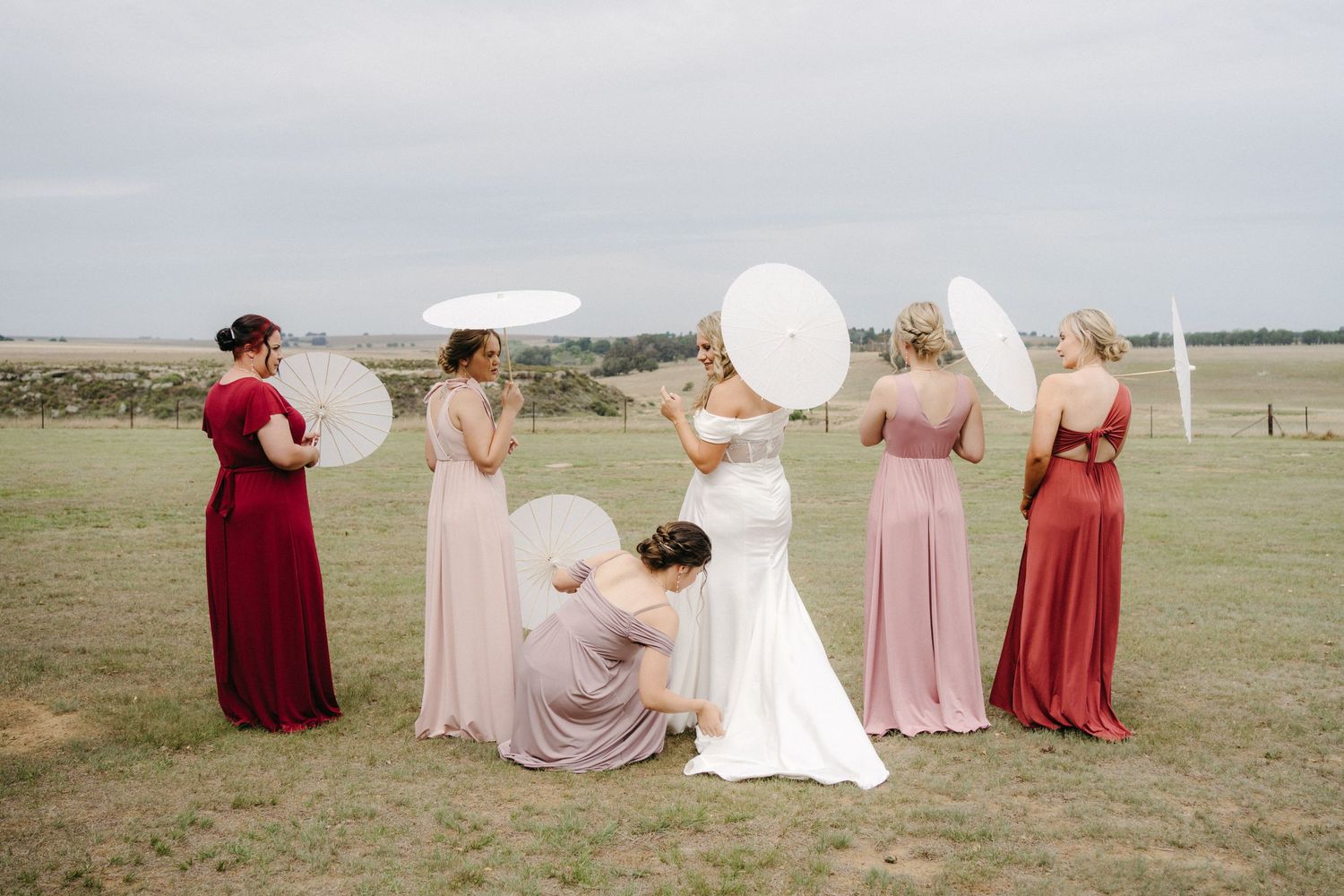 Wedding party poses with parasols in an outdoor field wearing pink and burgundy dresses.