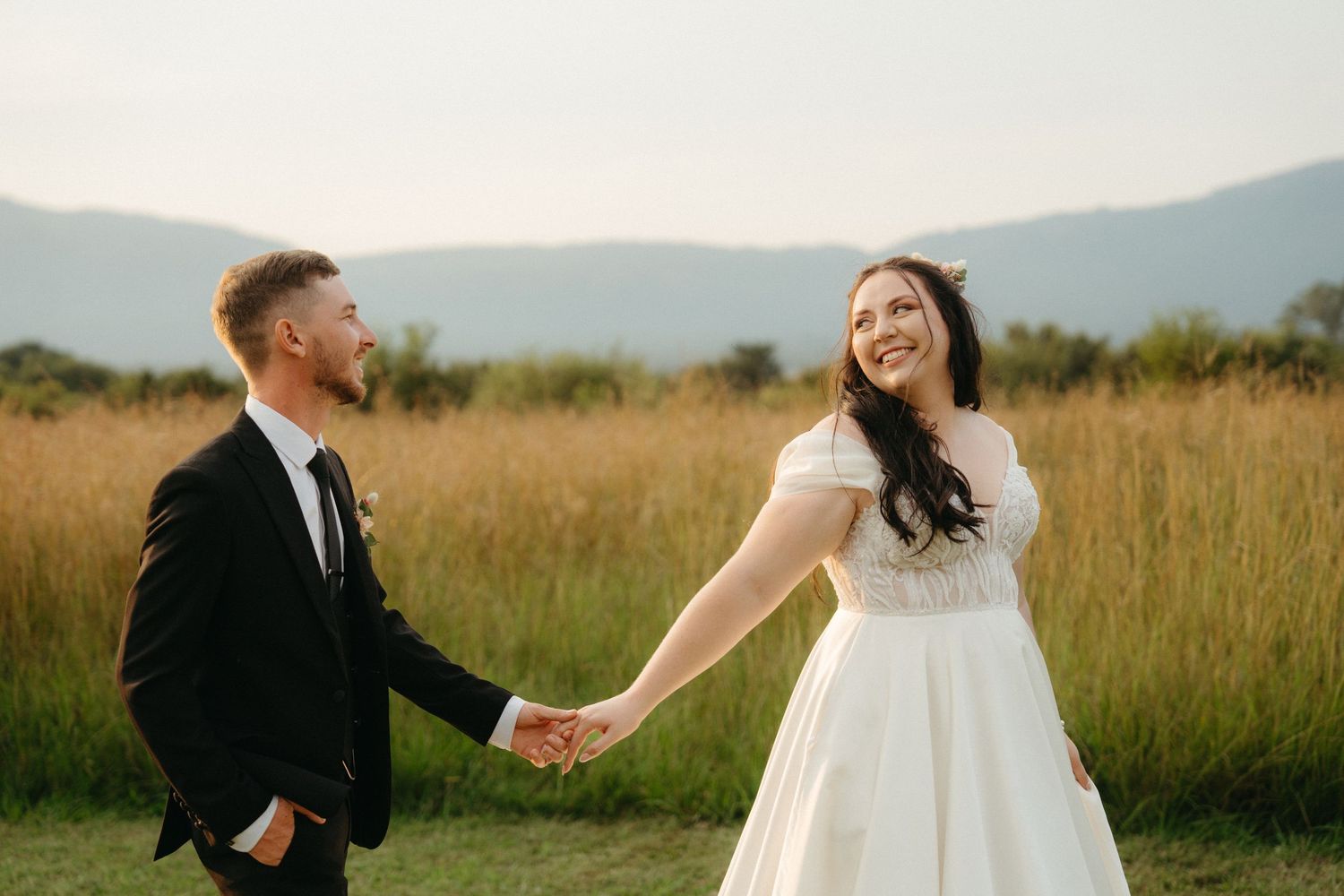 Joyful couple laughs while holding hands in meadow with mountain backdrop.