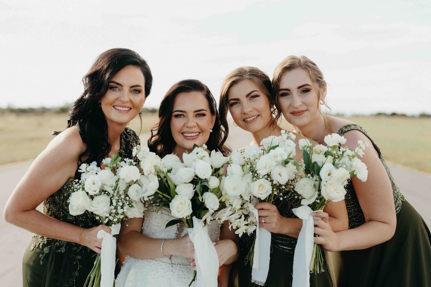 Bride and three bridesmaids in olive green dresses holding white rose bouquets, smiling outdoors on a sunny day.