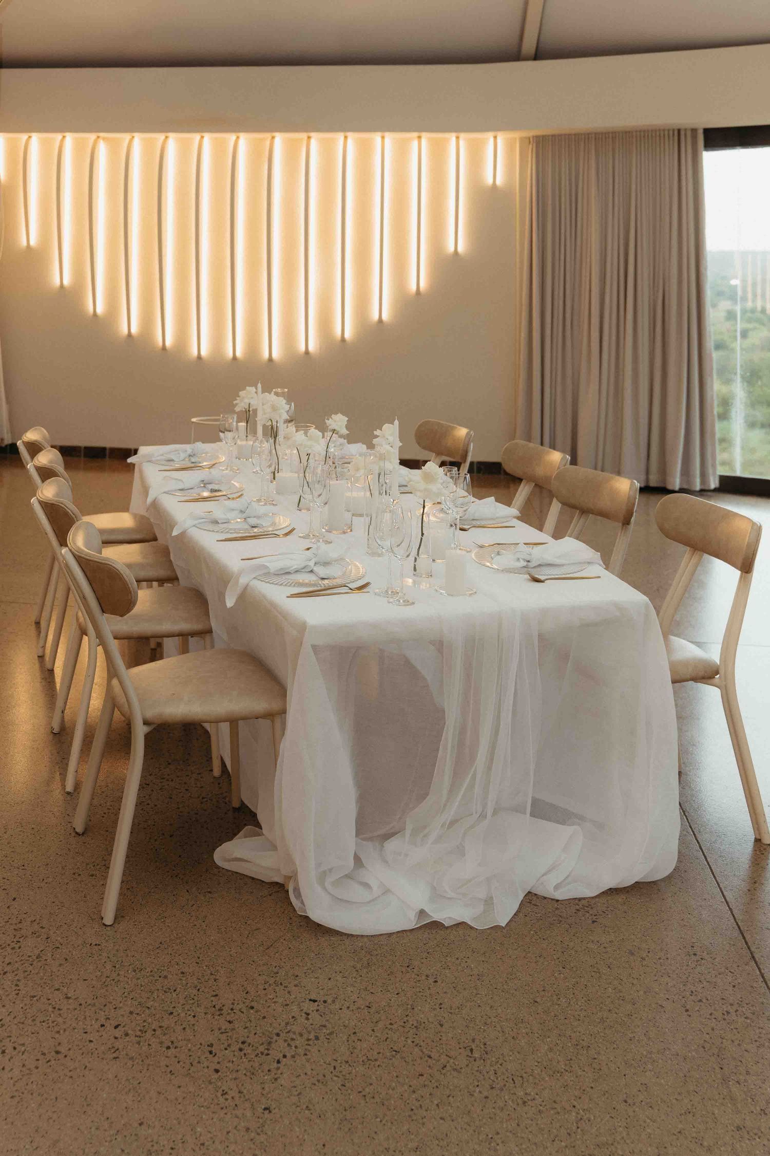 Elegant white tablecloth dining table set for a wedding reception with neutral chairs and warm glowing wall backdrop.