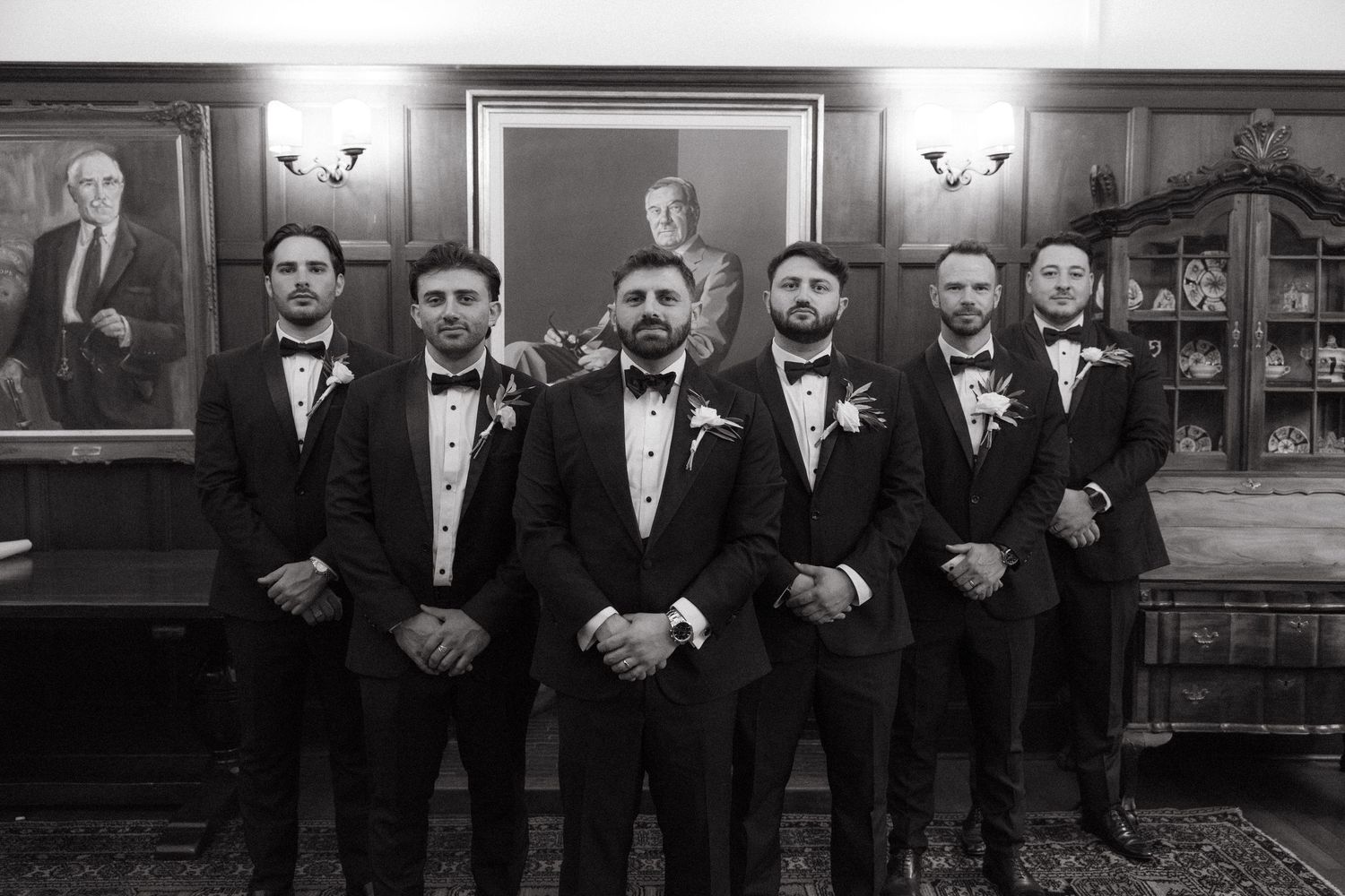 Groom and groomsmen in black tuxedos with bow ties posing formally in black and white photo indoors.