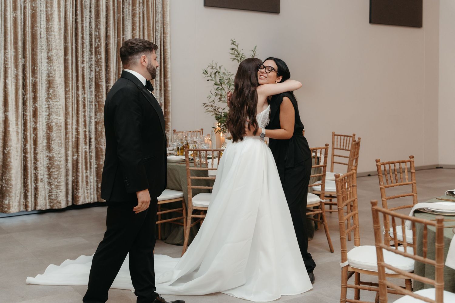 Bride in white gown hugging guest in black dress while groom watches at elegant wedding reception venue.