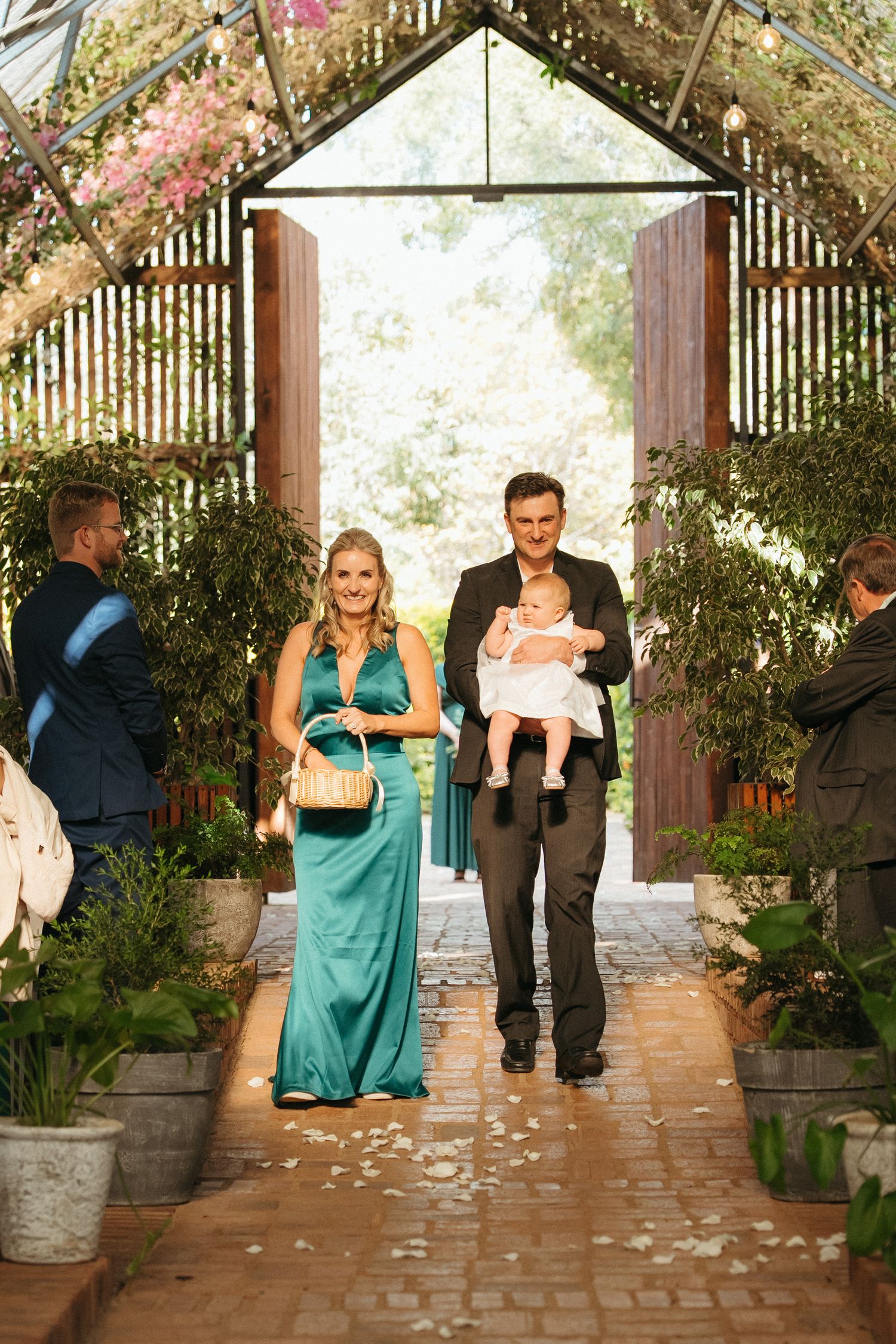 A couple in formal attire walks down a flower-lined aisle, the man holding a baby, at an elegant greenhouse wedding.