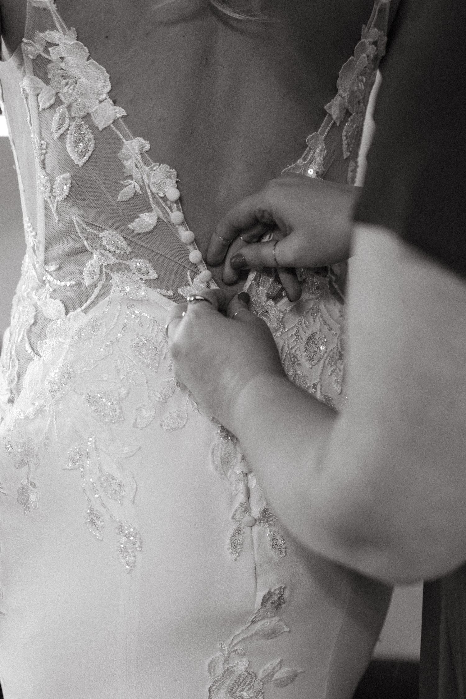 Black and white photo of hands fastening buttons on a lace wedding dress.
