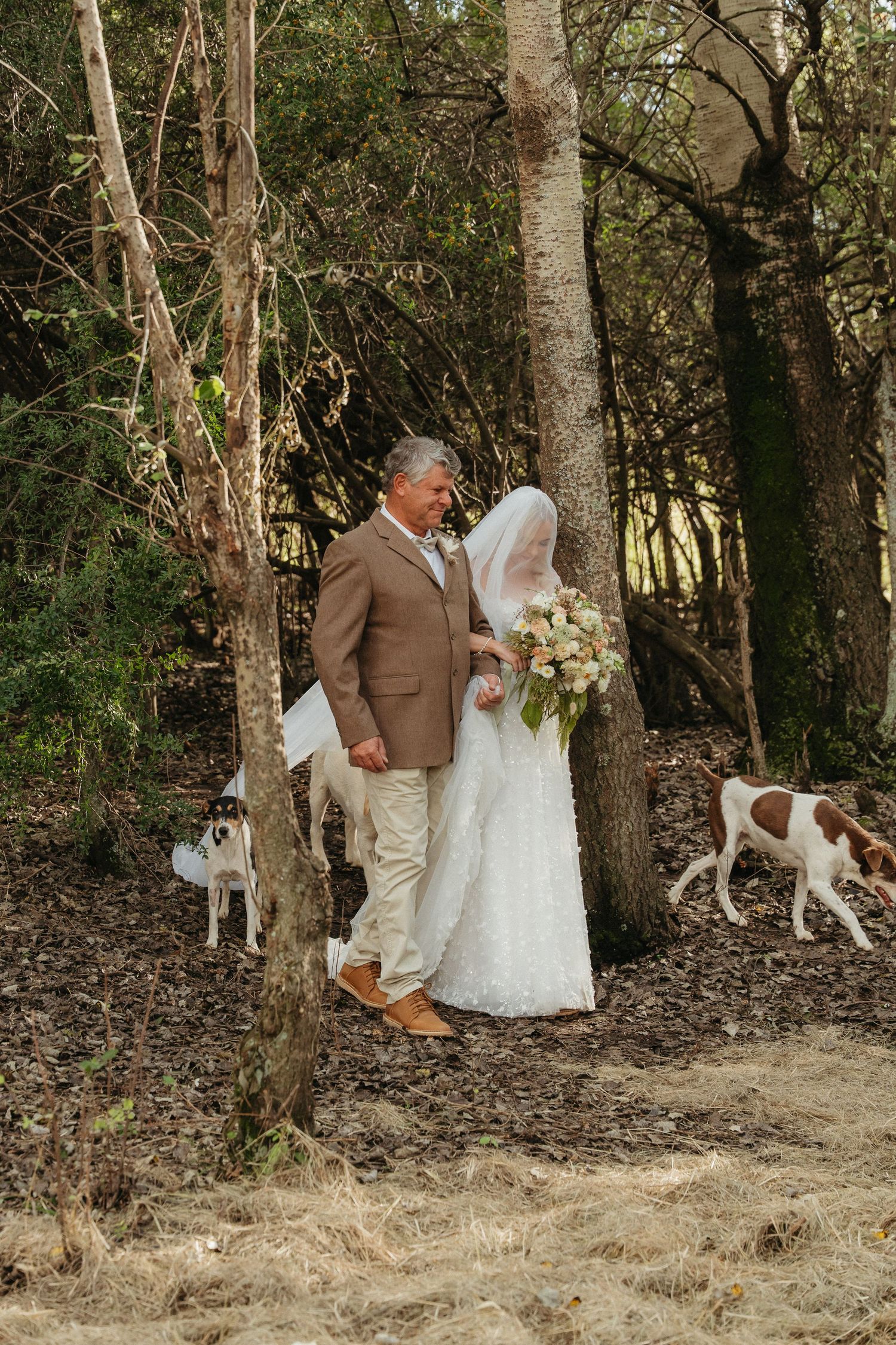 Bride in white lace gown holding bouquet walks with man in brown suit through wooded area as dogs roam nearby.