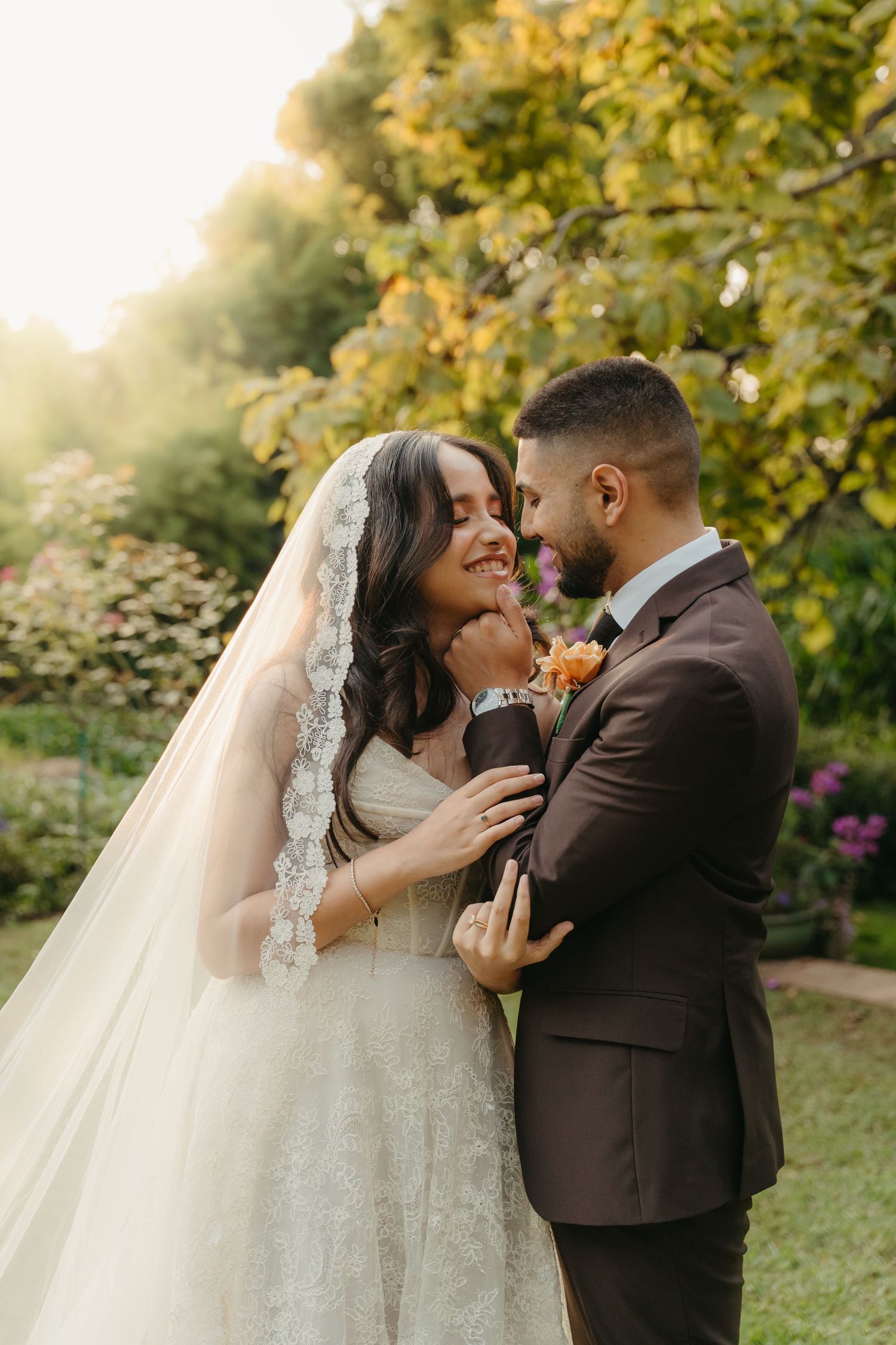 Bride and groom embrace in a sunlit garden, she wears lace veil and gown, he wears a dark brown suit with floral boutonniere.