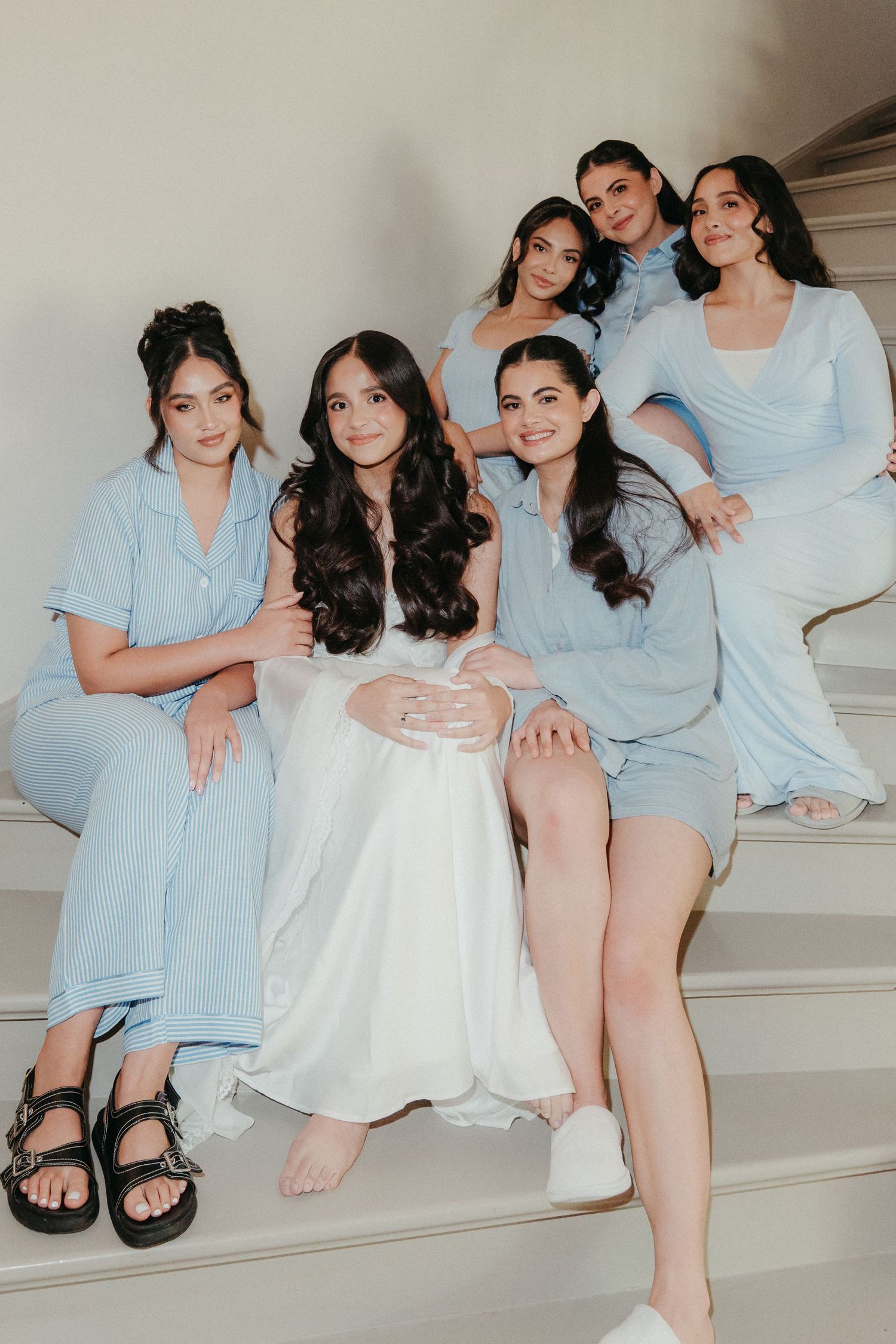 Six women in light blue and white outfits pose together on elegant white stairs for a bridal party photo.