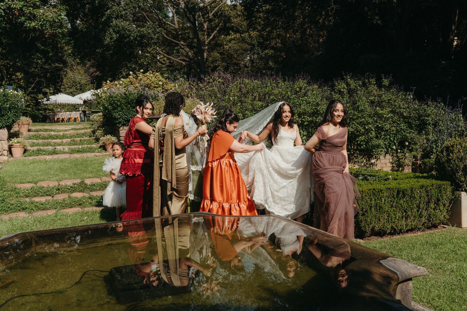 Bride in white gown with veil held by bridesmaids in colorful dresses beside a garden pond at Beechwood gardens