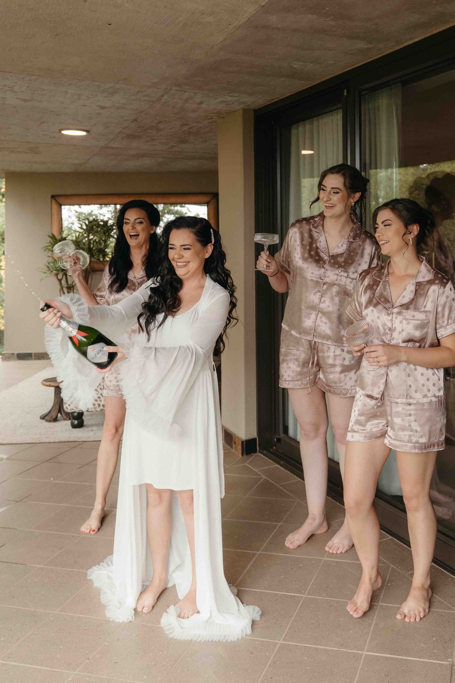 Bride in white robe pops champagne with bridesmaids in satin pajamas on a patio.