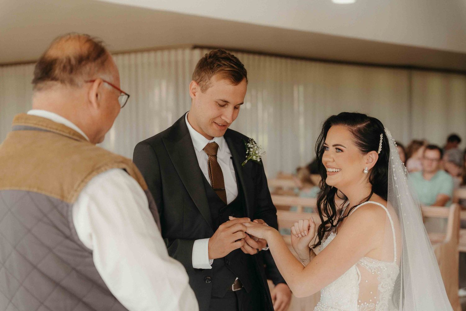 Bride and groom exchanging rings during wedding ceremony with officiant, bride smiling joyfully in white gown.