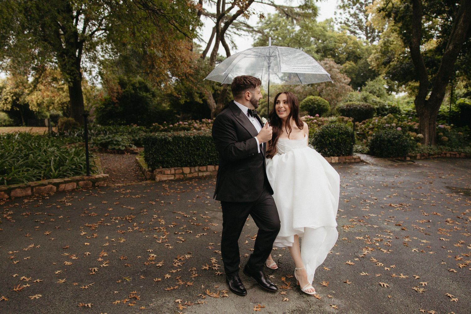 Bride and groom share a romantic rainy day wedding photo under a clear umbrella surrounded by autumn foliage.