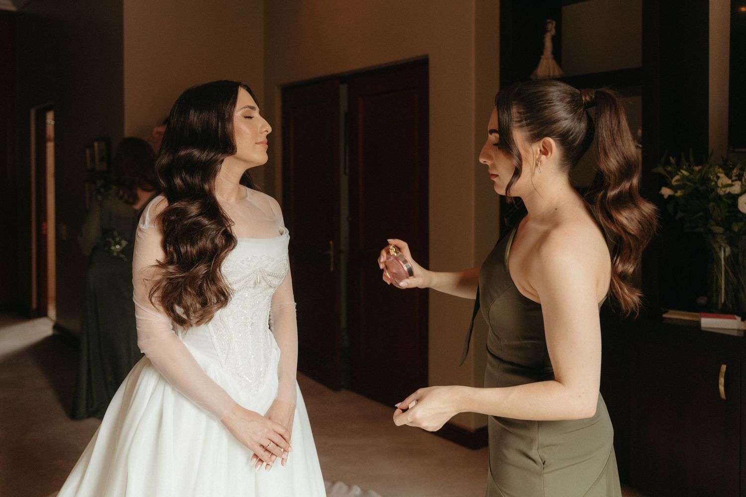 Bride in white gown getting ready with bridesmaid in olive dress helping with final touches indoors.