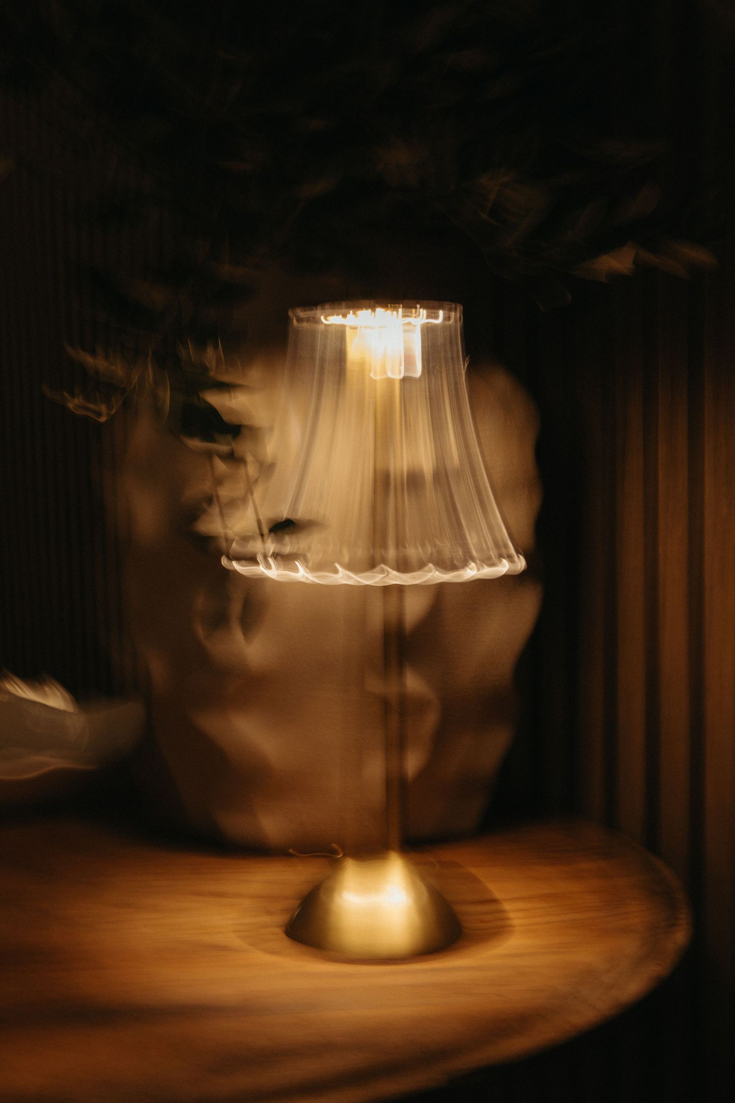 A glowing table lamp with a translucent shade on a wooden table, casting warm light in a dark room.