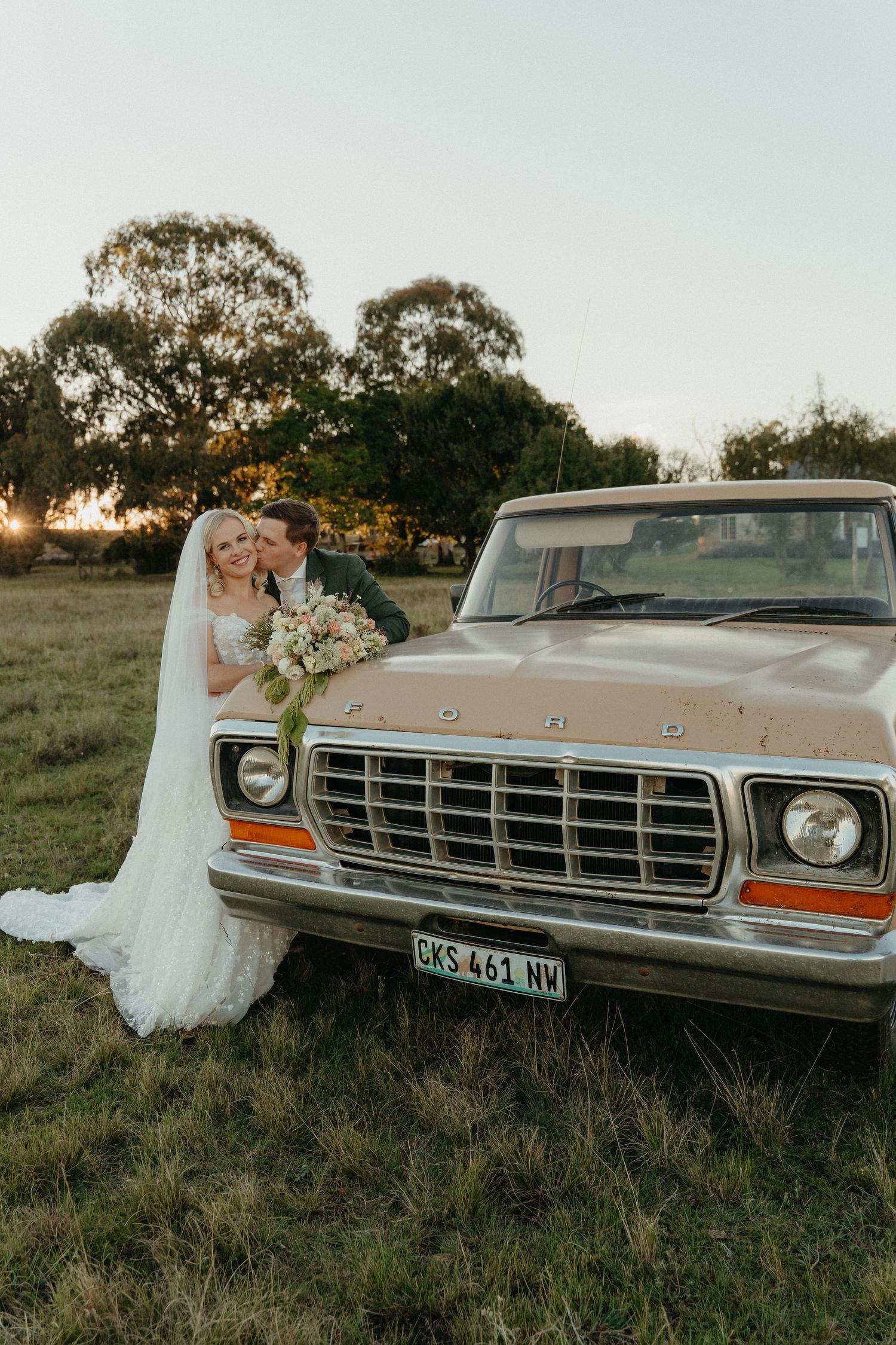 Bride in a lace gown and veil holding a bouquet kisses the groom beside a vintage Ford pickup truck in a grassy field at sunset.