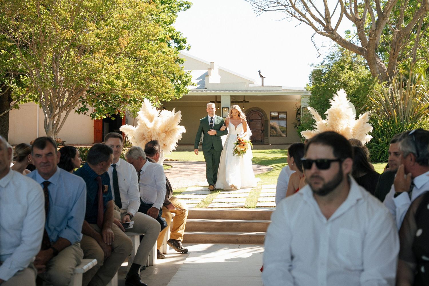 Father walks bride in white gown down the outdoor aisle past seated wedding guests and pampas decorations during a sunny ceremony at Duvon.