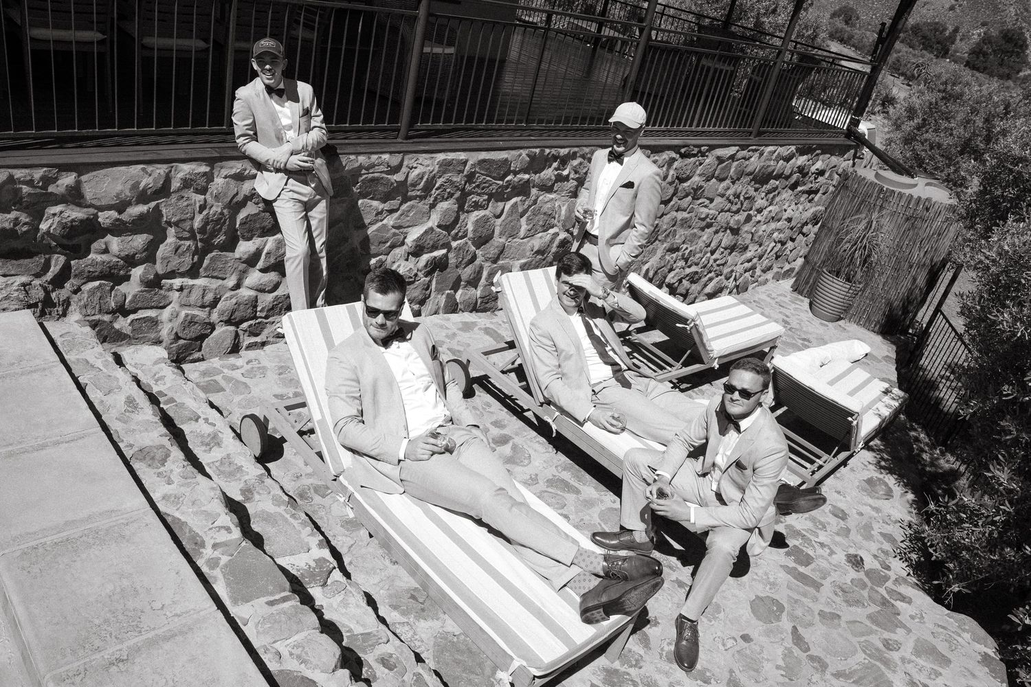 Black and white photo of four men in suits relaxing on lounge chairs and standing on a stone patio beside a rustic stone wall.