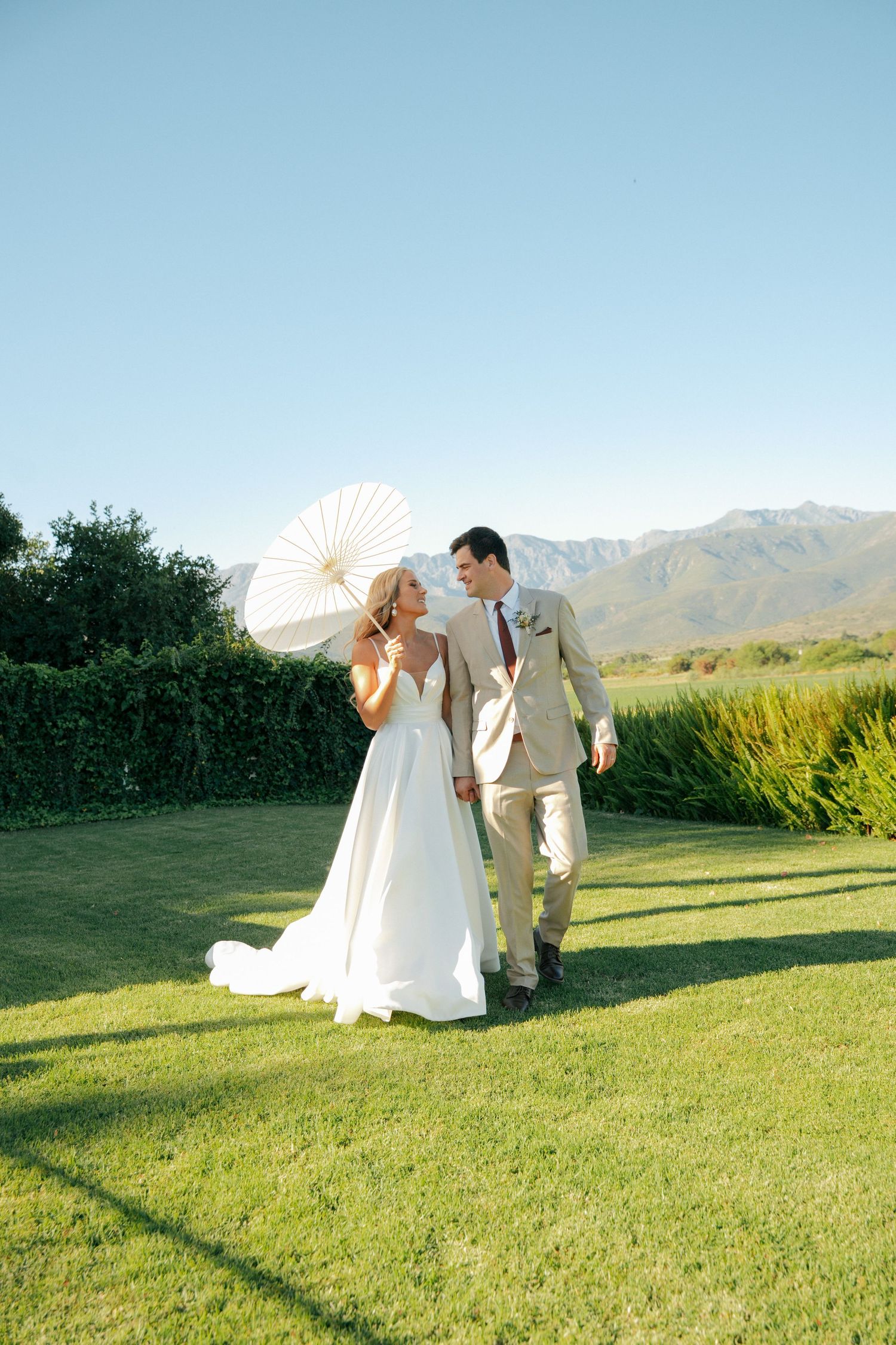 A bride in a white gown holding a white parasol walks hand in hand with a groom in a tan suit across a sunny lawn at Duvon Wine Estate