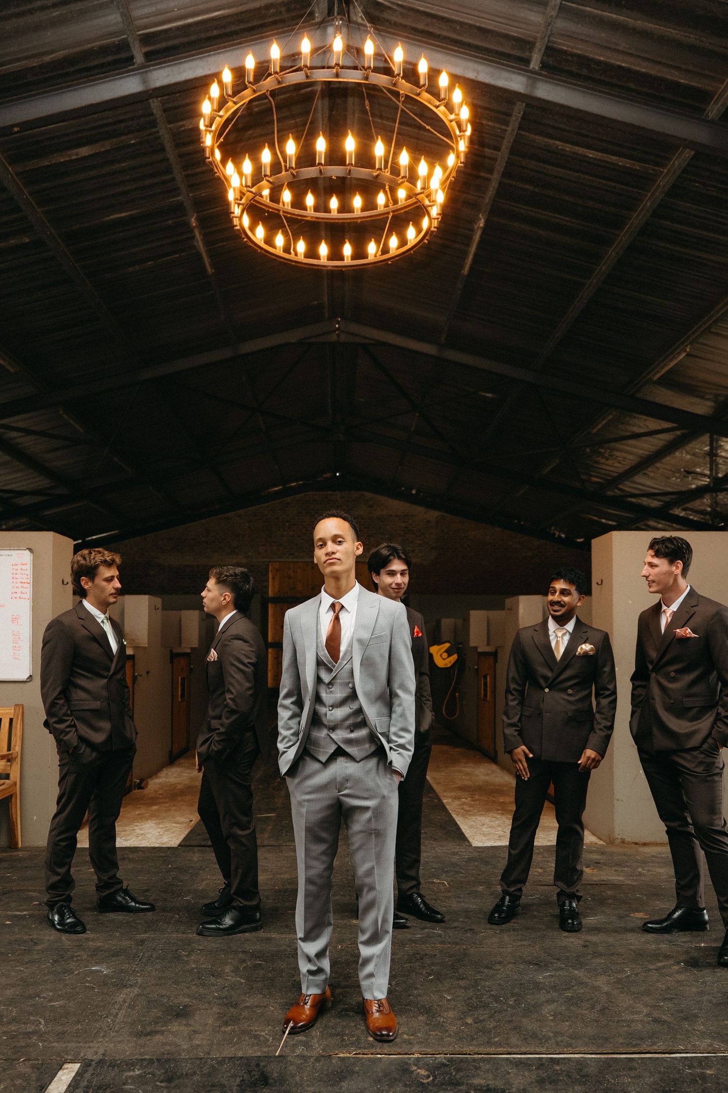 Groom in light gray suit stands centered before groomsmen in brown suits under rustic chandelier in the stables at Tafika Polo Farm.