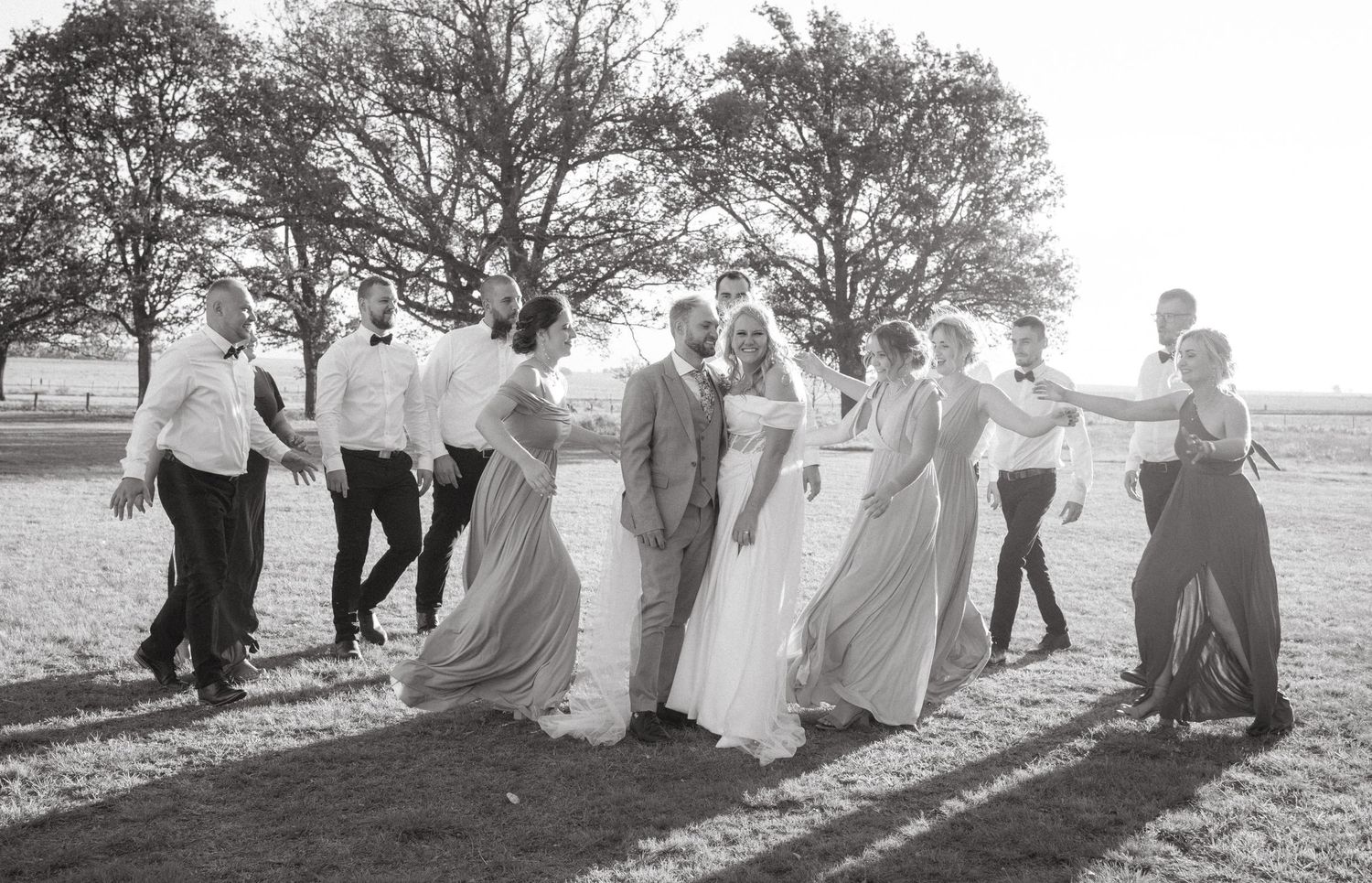 A wedding party walks together outdoors in a playful candid moment, with long dresses flowing in black and white.